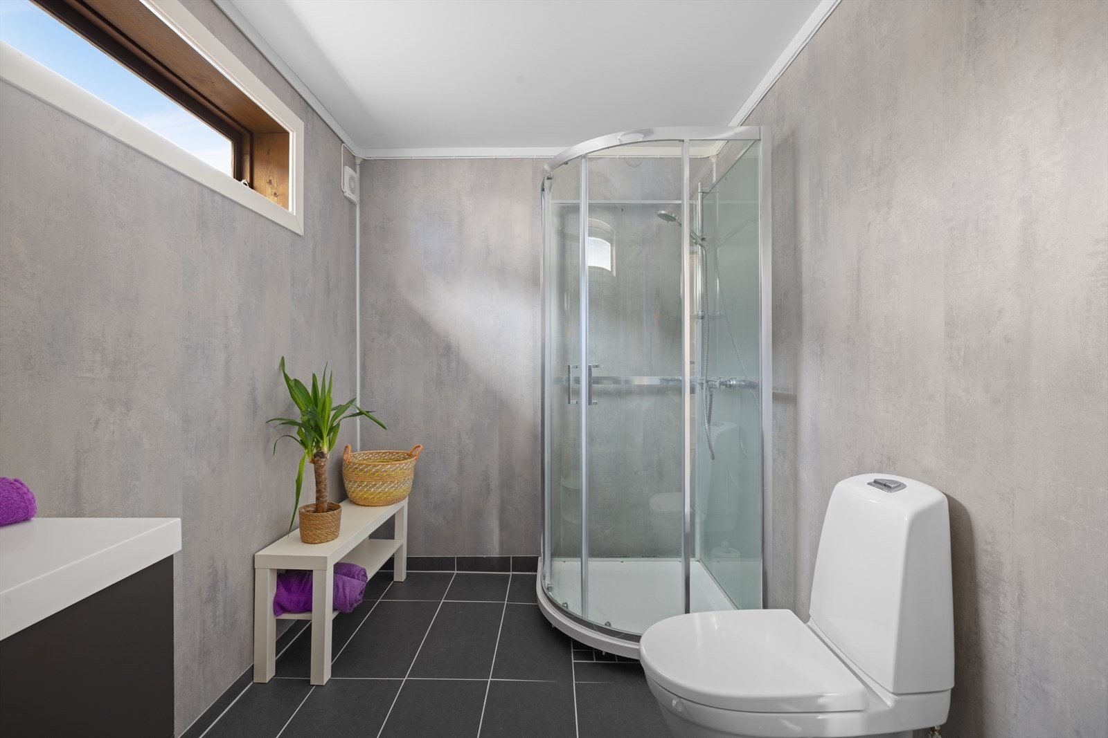 Badet har fliser på gulv med varmekabler og våtromsplater på vegger. Rommet er utstyrt med heldekkende servant over skuffeinnredning, speil, toalett og dusjkabinett Galleribilde