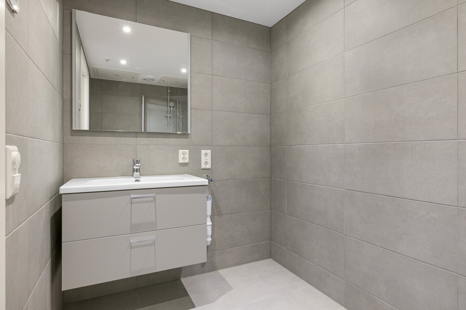 Badet er lekkert med store fliser på vegger og gulv. Badet har downlights og vannbåren gulvvarme. Den øvrige innredningen består av servant med skuffer under og et stort speil på veggen. Galleribilde