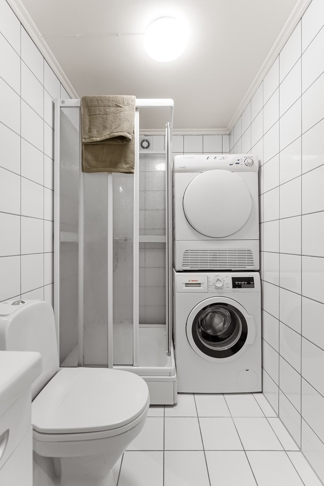 Badet har toalett, vegghengt servantskap med ovenpåliggende servant, speil, overskap og lys over servant, dusjkabinett og opplegg for vaskemaskin Galleribilde