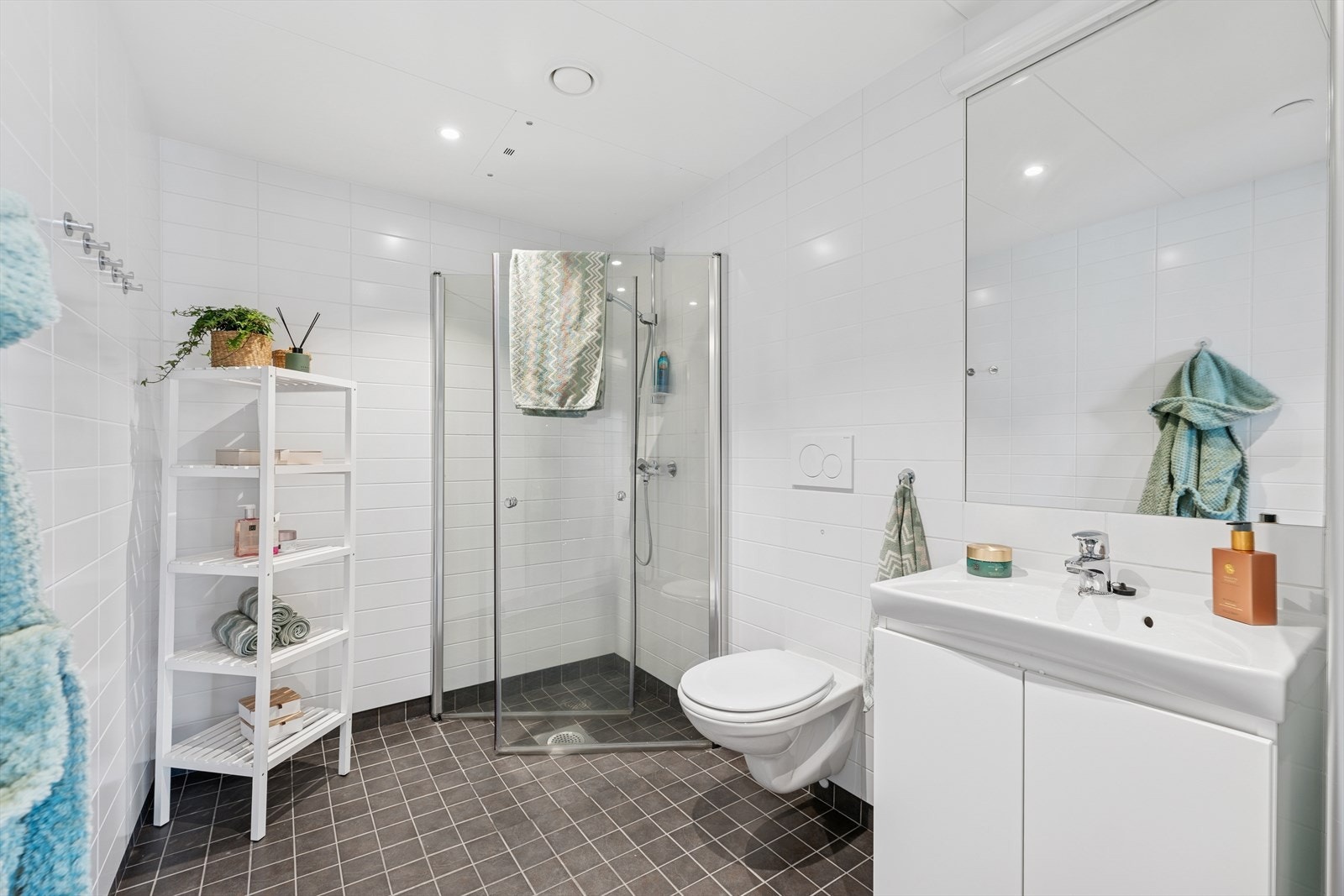 Badet er innredet med heldekkende servant på benkeskap, speil med lys, vegghengt toalett, dusj i hjørnet bak svingbare glassvegger og opplegg for vaskemaskin Galleribilde