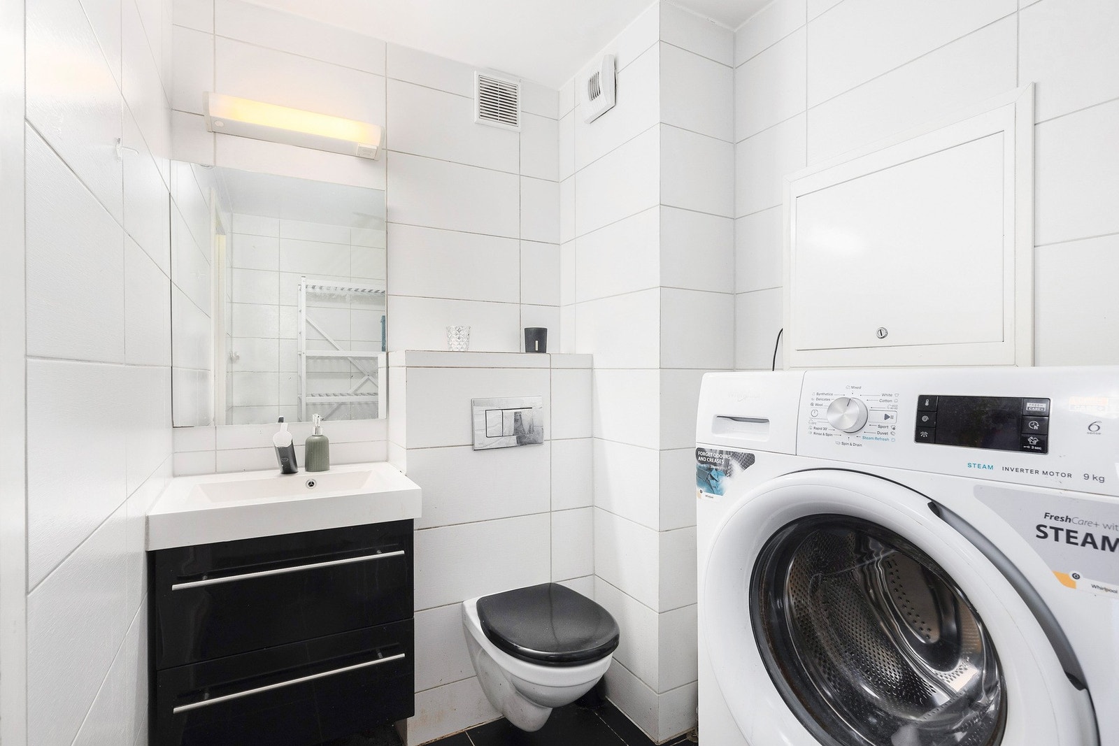 Badet har hvite fliser på veggene og flislagt gulv. Innredningen består blant annet av nedfelt servant, vegghengt toalett og opplegg for vaskemaskin. Galleribilde