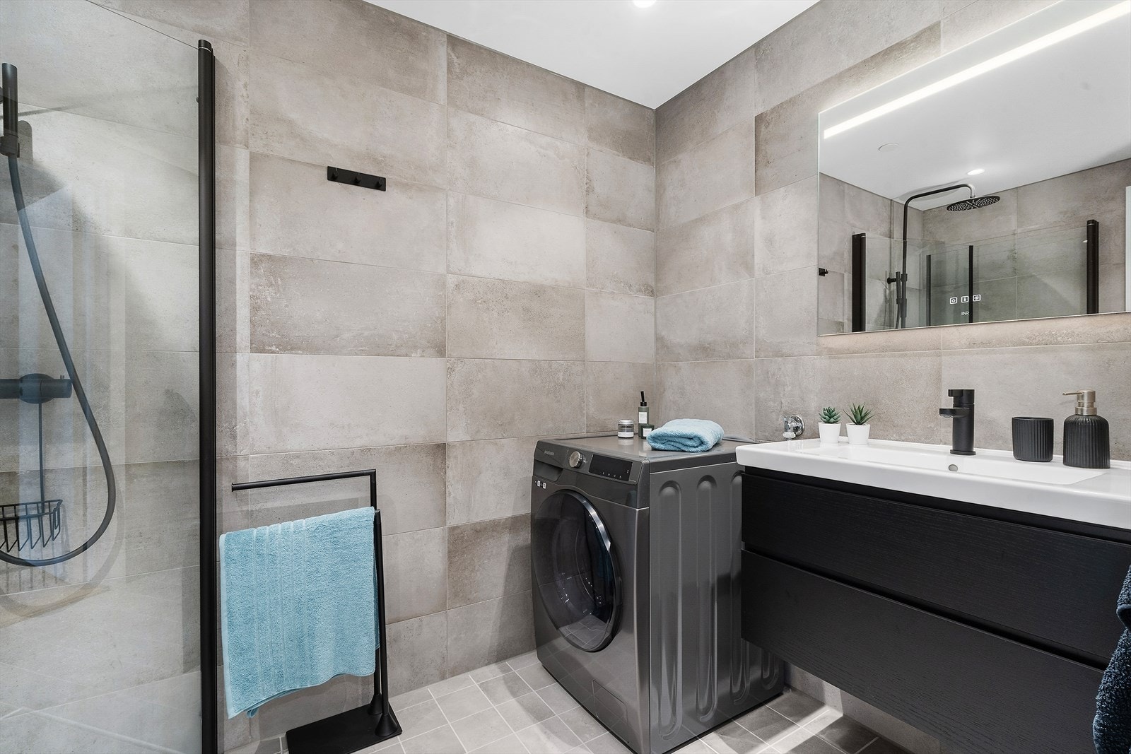 Tidløst innredet, flislagt bad med varmekabler i gulv. Badet er utstyrt med sort baderomsinnredning med heldekkende nedfelt servant, speil med belysning, vegghengt toalett og dusjhjørne med innfellbare dusjvegger. Galleribilde