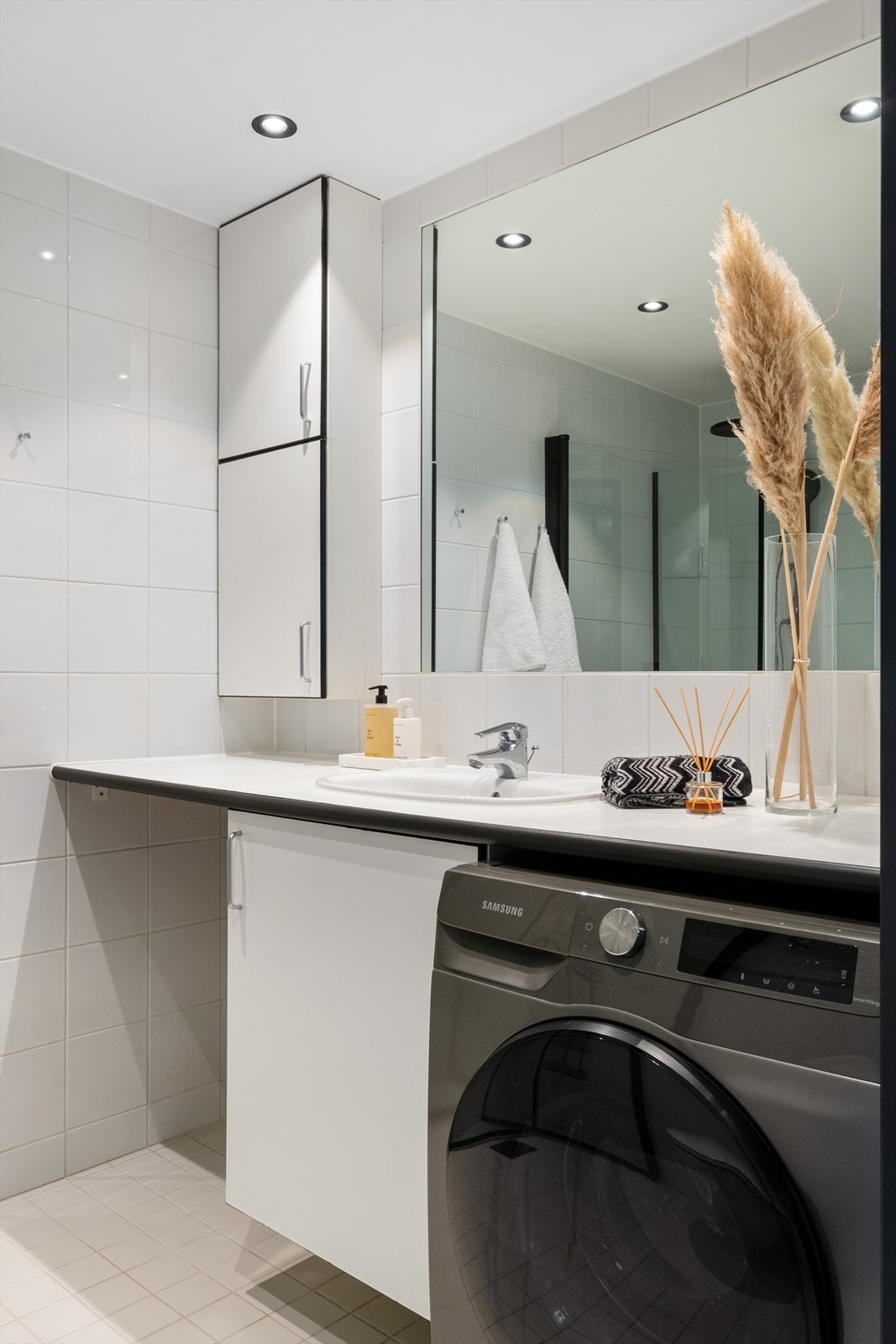 Baderomsinnredning med lyse flater, stort innfelt speil og opplegg for vaskemaskin og tørketrommel. Galleribilde
