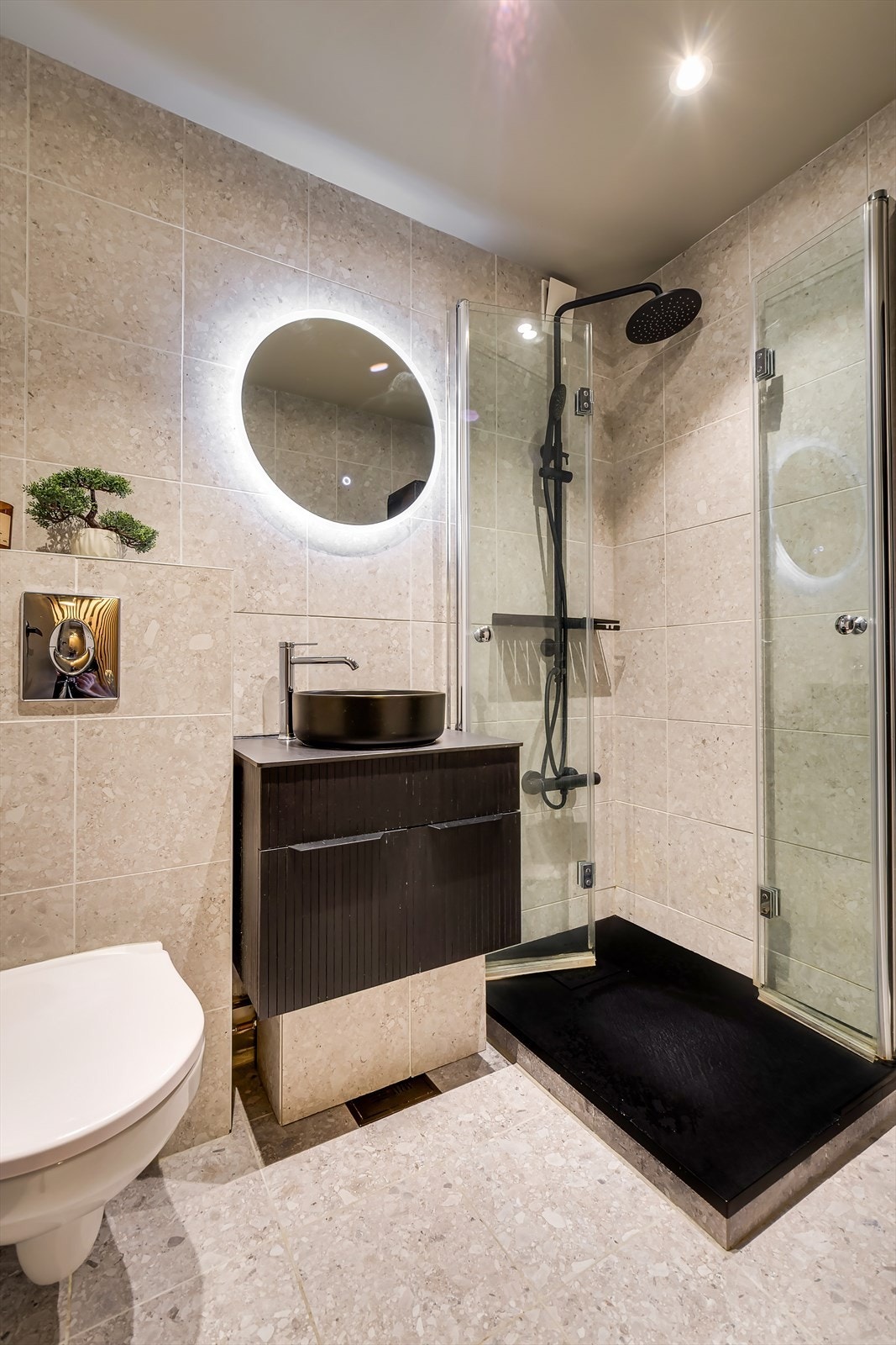 Badet har et moderne utseende og en praktisk utforming med dusj med svingbare glassdører, veggmontert toalett med innebygget sisterne, servant med underskap, høyskap og speil med integrert belysning. Galleribilde