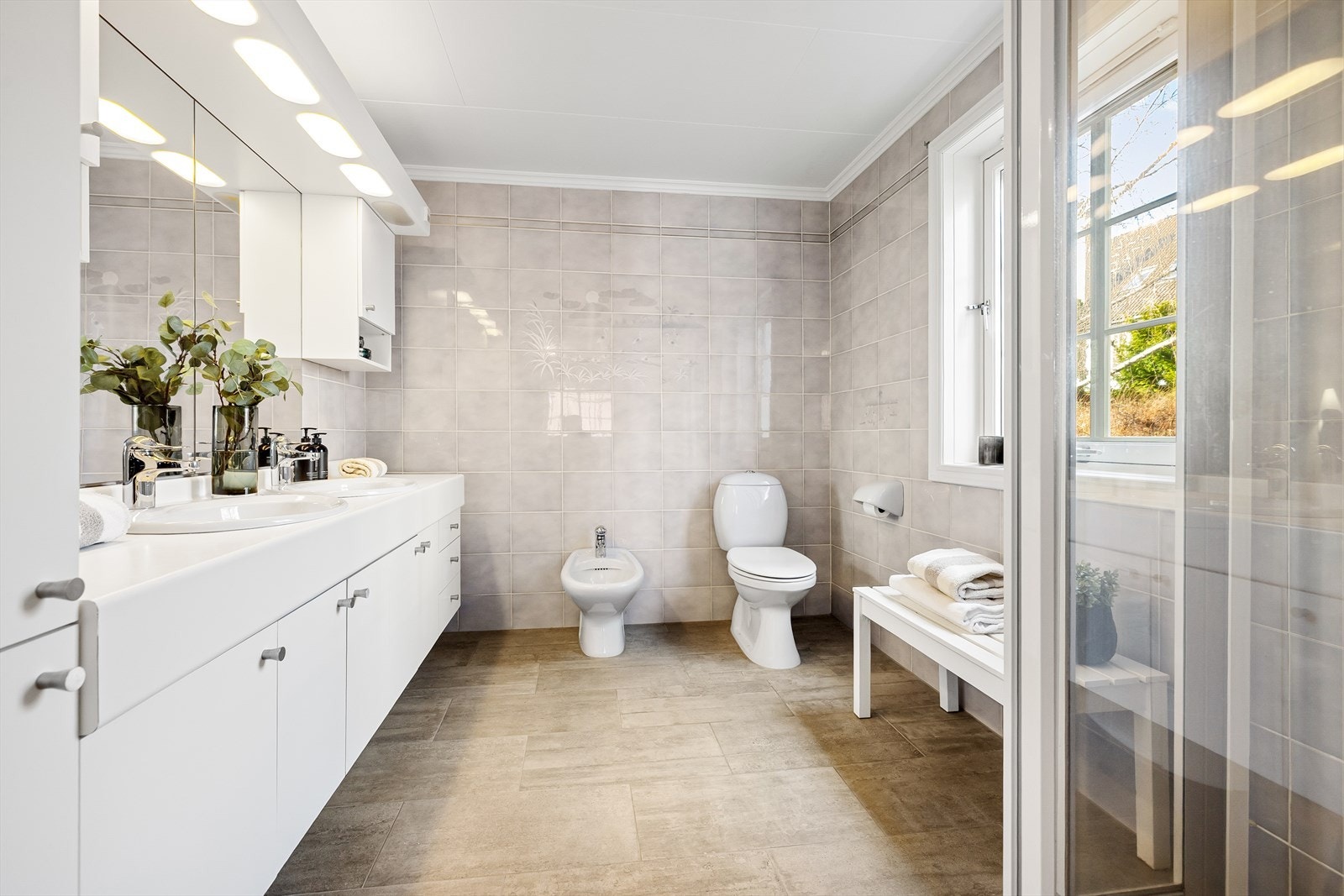 Romslig bad med varmekabler i gulv og moderniseringer i form av flis på flis på gulv. Her er det 20 x 25 cm fliser på vegger og 60 x 30 cm fliser på gulvet samt 5 x 5 cm fliser i dusjsonen. Belysningen kommer fra lys over speil og fra vinduet. Galleribilde