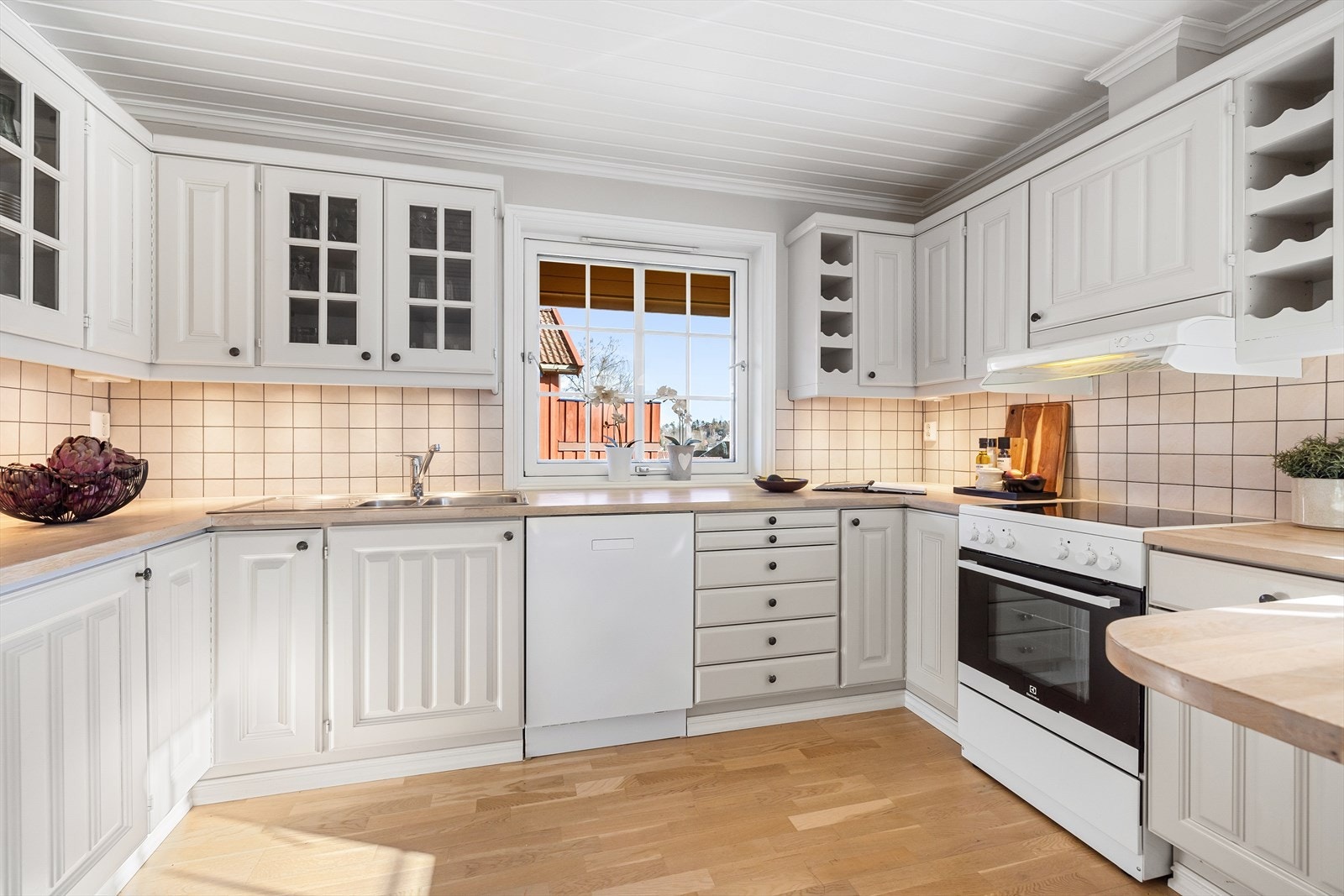 Kjøkkeninnredning m/profilerte fronter, 2 integrerte vinhyller og overskap, hvor noen fronter har glass. Benkeplate av heltre med en metallvask, og det er fliser mellom benken og overskapene. Belysning under overskapene og lamper som lyskilde. Galleribilde