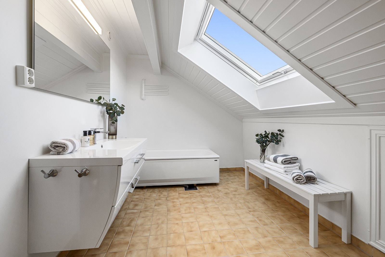 Flott bad med varmekabler i gulv. Her er det 15 x 15 cm fliser på gulvet og vegger er slette og malte. Badet har skråtak med et Velux-vindu, som sørger for lysinnslipp. Galleribilde