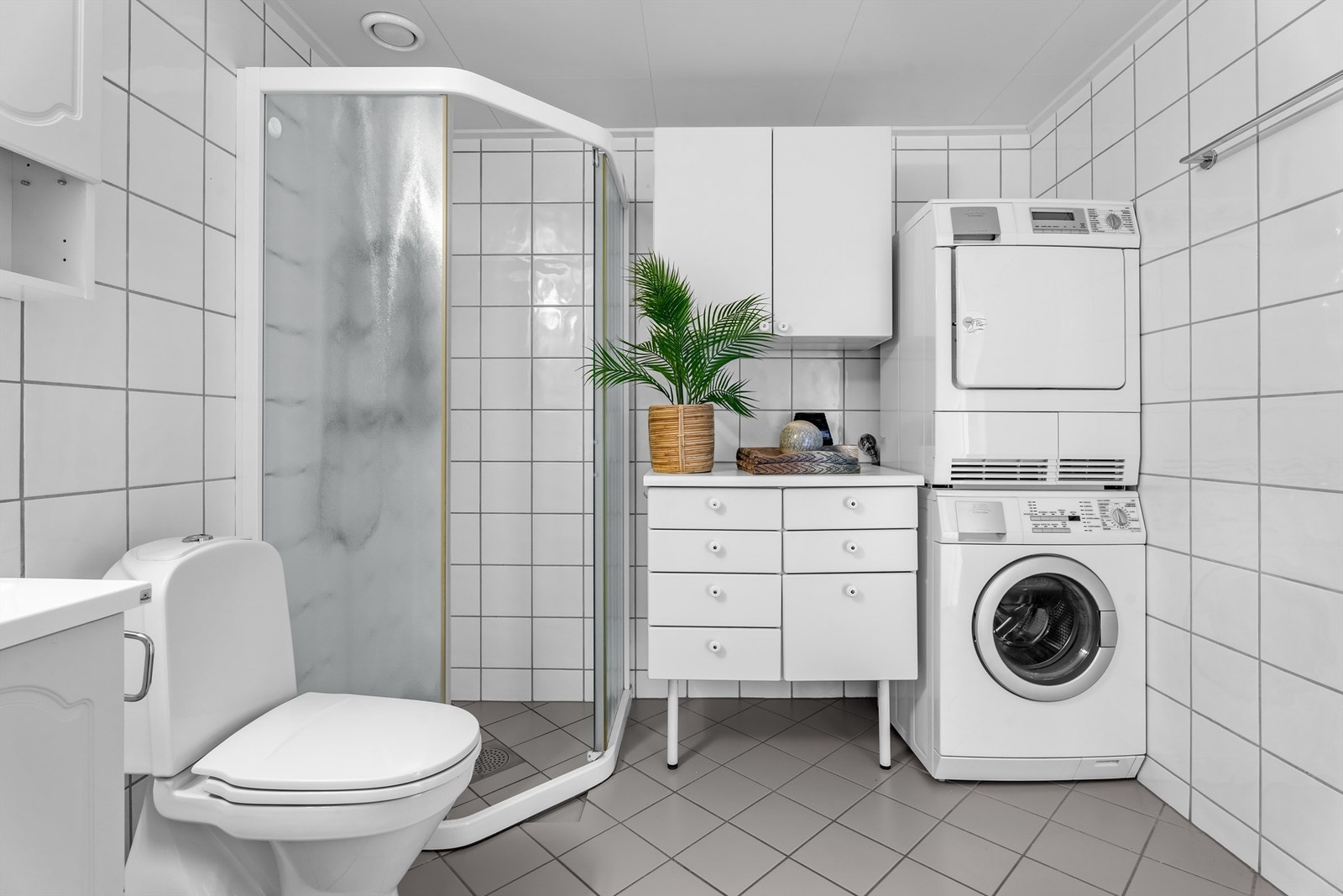 Baderommet er utstyrt med vegghengt servantinnredning, gulvstående baderomsinnredning og overskap, dusjhjørne med glassfelt og skyvedør av glass, toalett og opplegg for vaskemaskin og tørketrommel. Galleribilde