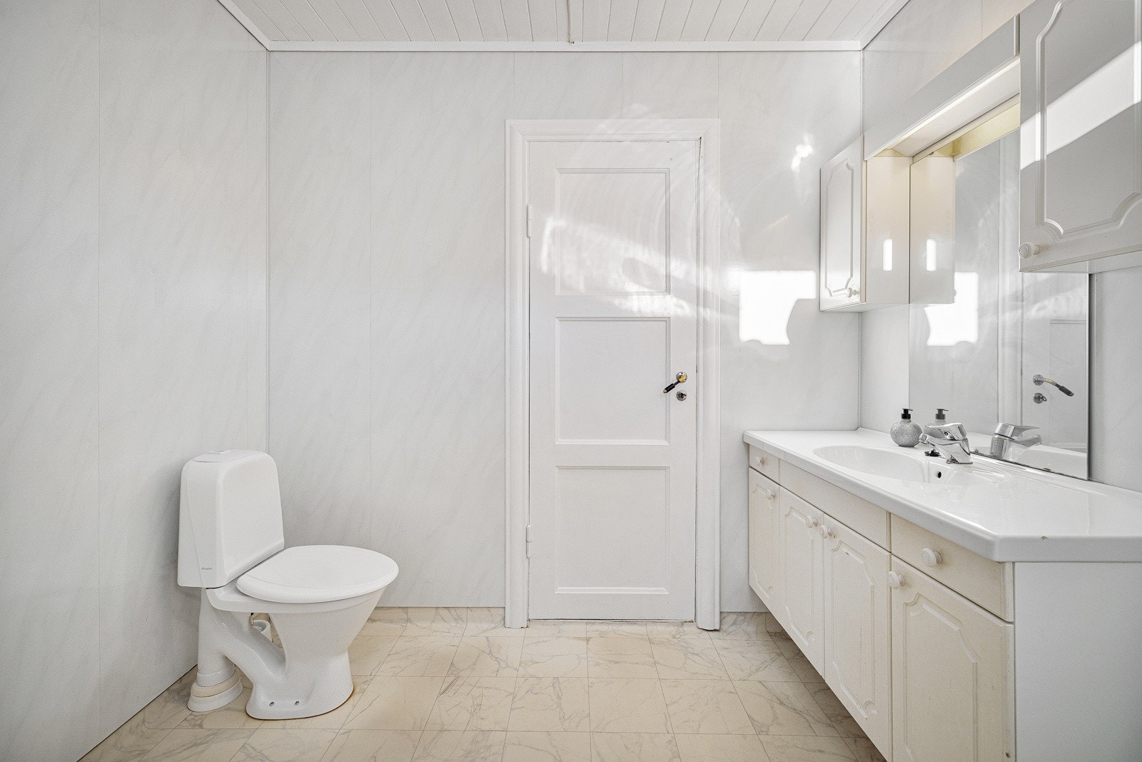 Badet er utstyrt med frittstående toalett, dusjhjørne og opplegg for vaskemaskin. Det er baderomsplater på vegger, vinylbelegg på gulv og himlingsplater i tak. Galleribilde