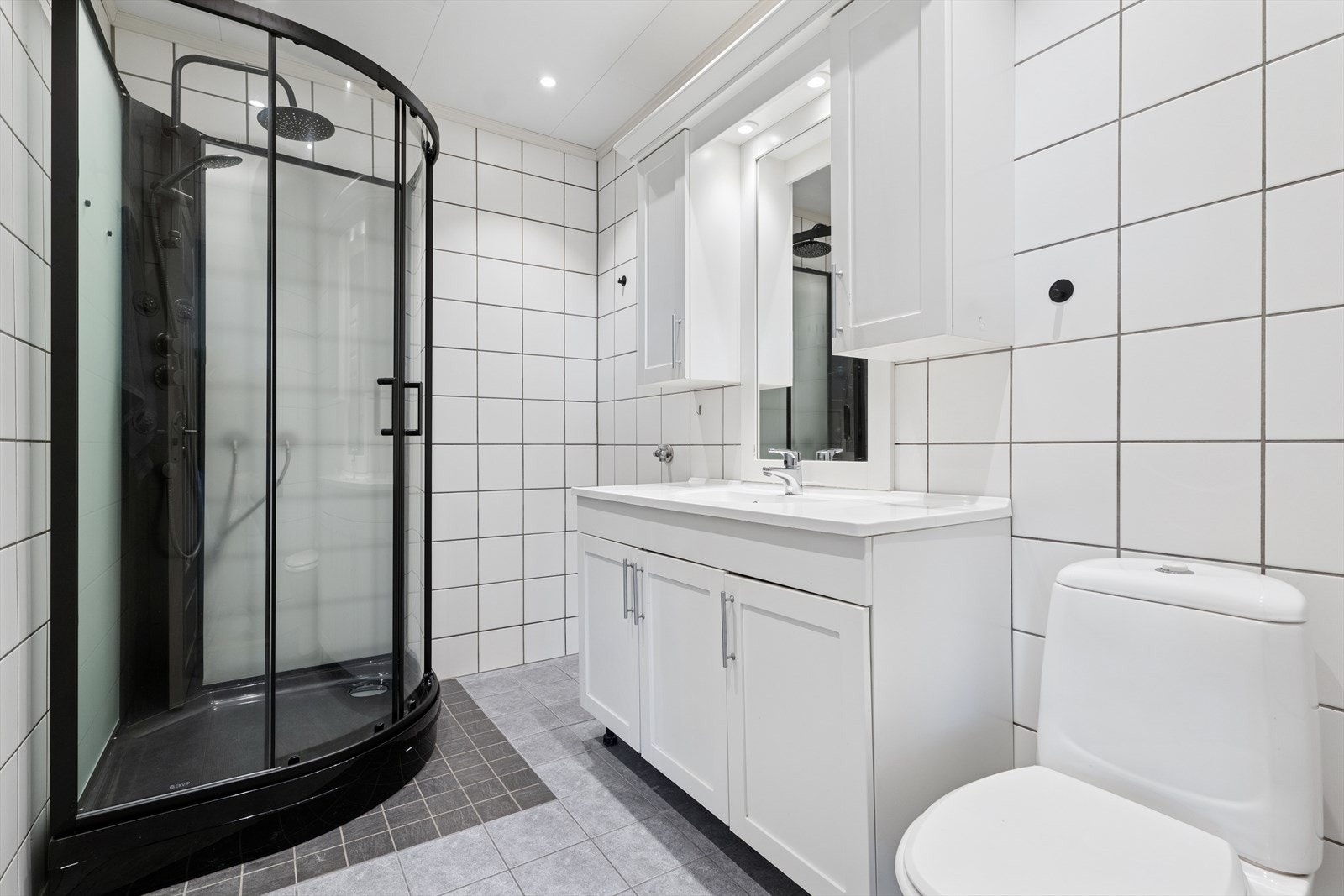 Lekkert baderom med fliser på gulv og vegger. Badet består av Dusjkabinett, toalett, servant med underskap, speil med tilhørende belysning og opplegg til vaskemaskin. Galleribilde