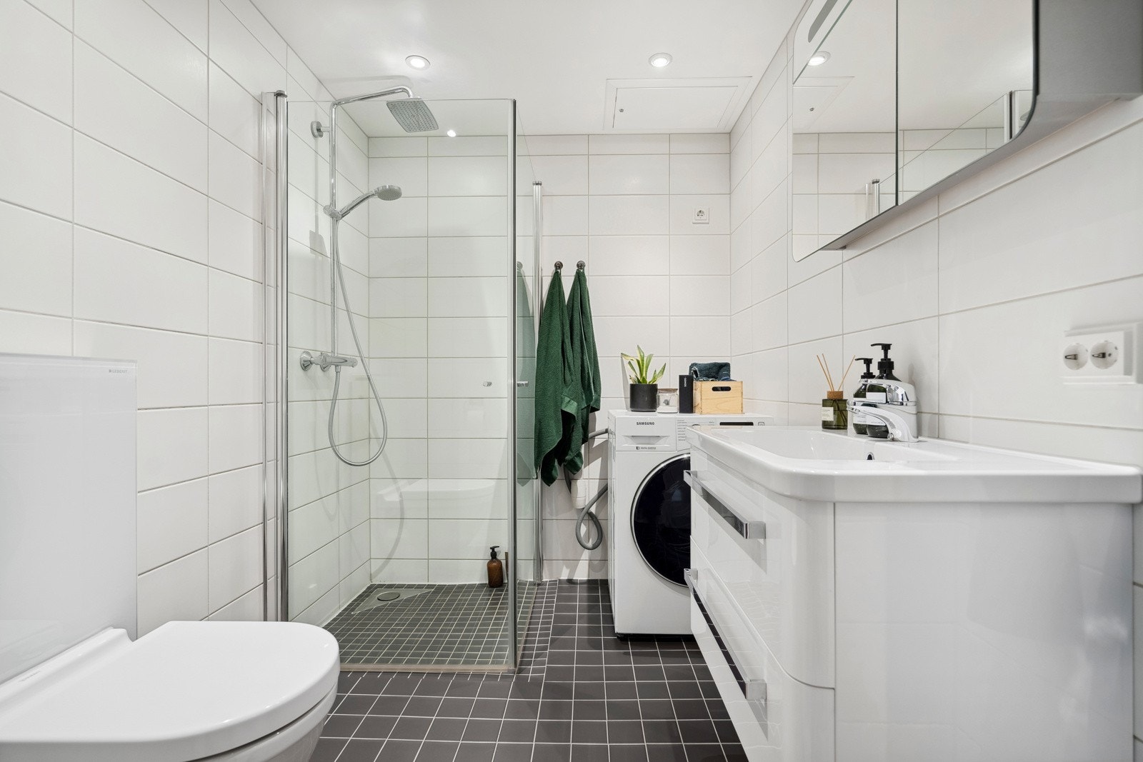 Flott og tidløst baderom innredet med servant, dusjvegger av glass og dusjgarnityr på vegg, samt opplegg for vaskemaskin. Galleribilde