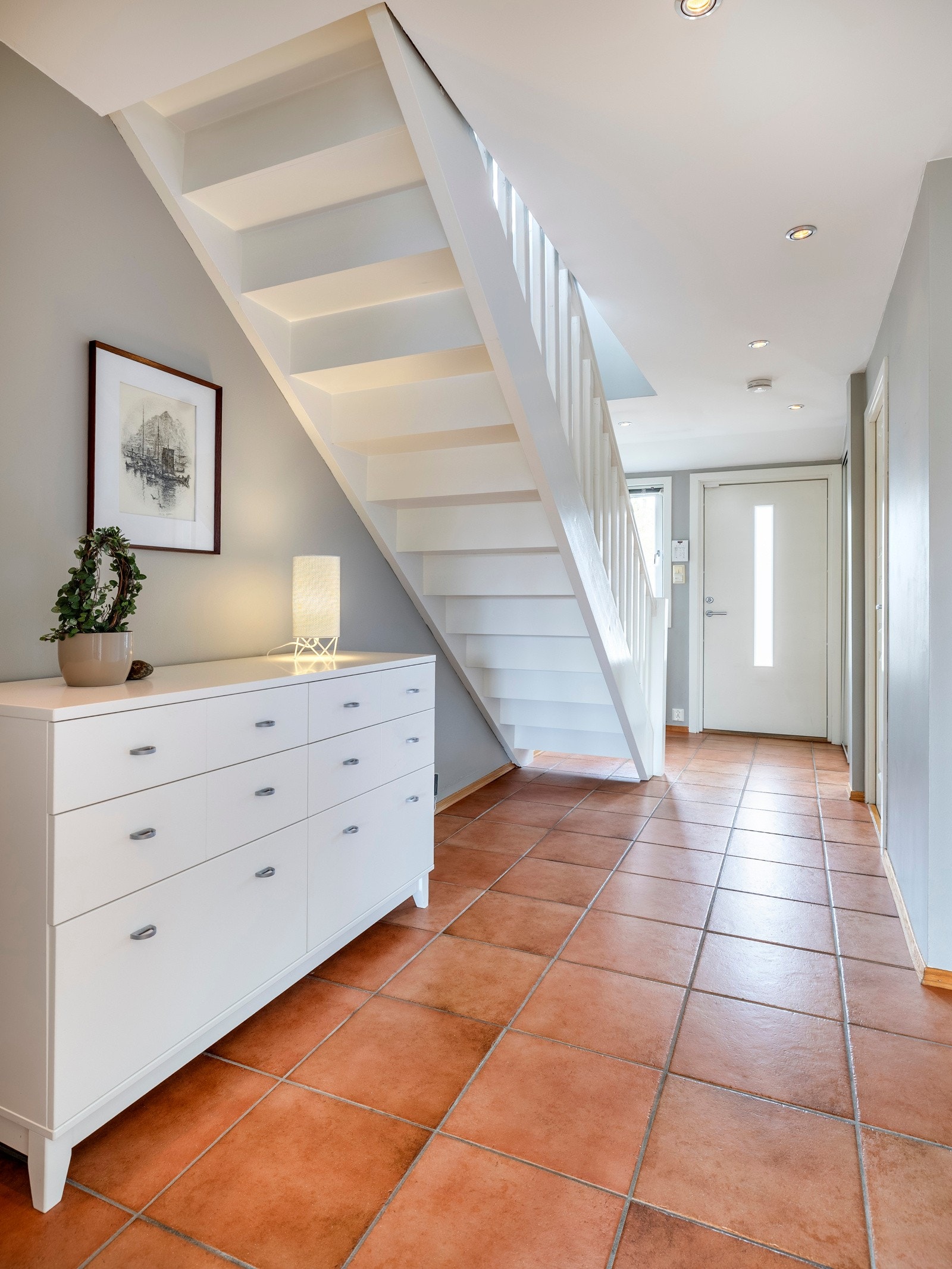 Entreen har terrakottafargede fliser på gulvet med varmekabler og en hvitmalt trapp med eiketrinn som fører opp til andre etasje. I den ene veggen er det innfelt en skyvedørsgarderobe med speilfronter. Galleribilde
