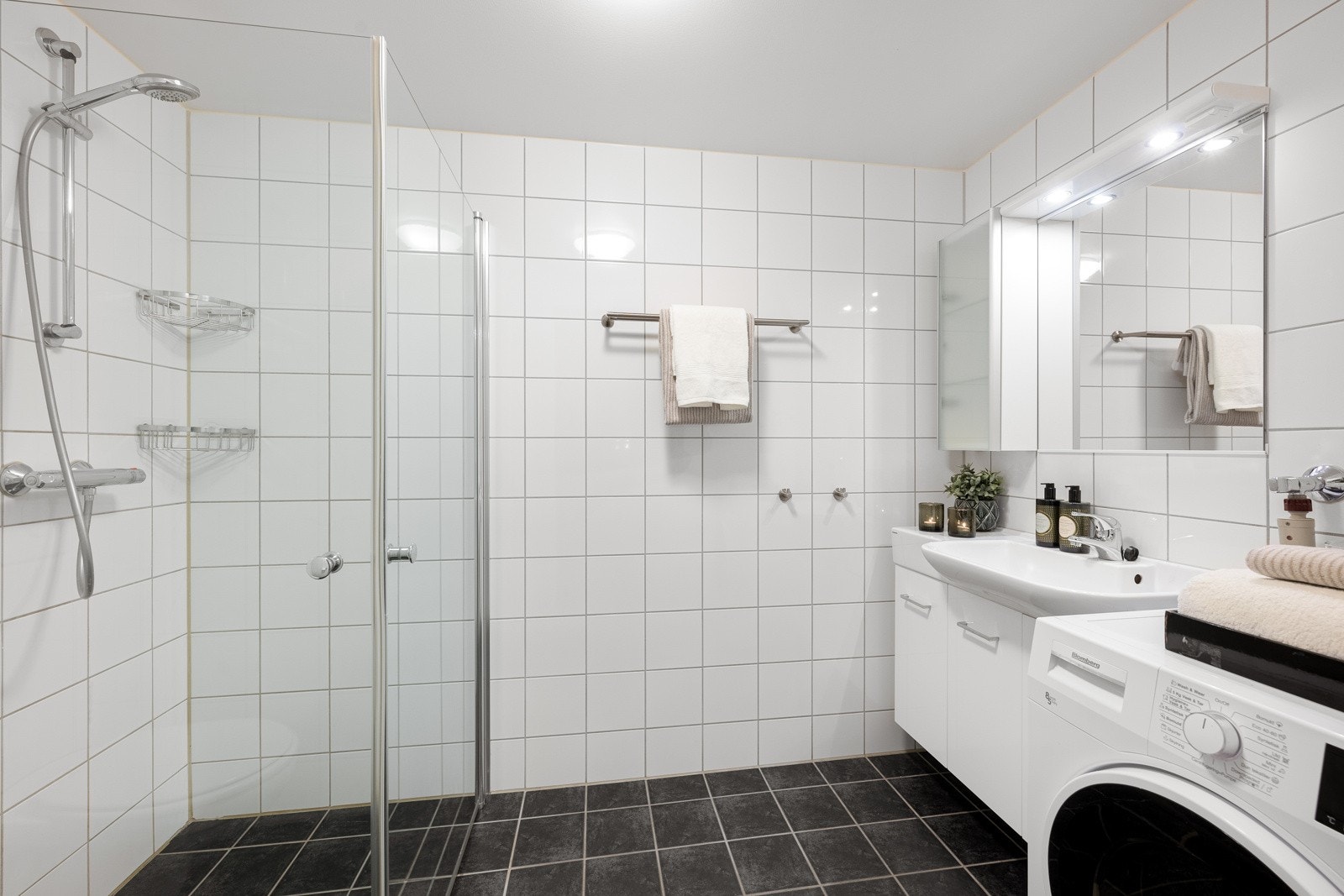 Moderne helfliset baderom med varmekabler i gulv. Badet inneholder: servant med møblement, speil og belysning, vegghengt wc, dusj og opplegg for vaskemaskin. Plass til tørketrommel Galleribilde