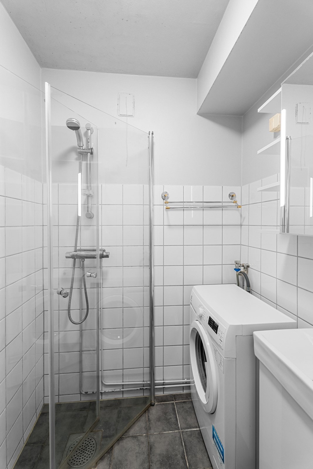 Badet har dusjhjørne med dører i herdet glass, gulvstående klosett, servant og opplegg for vaskemaskin. Galleribilde