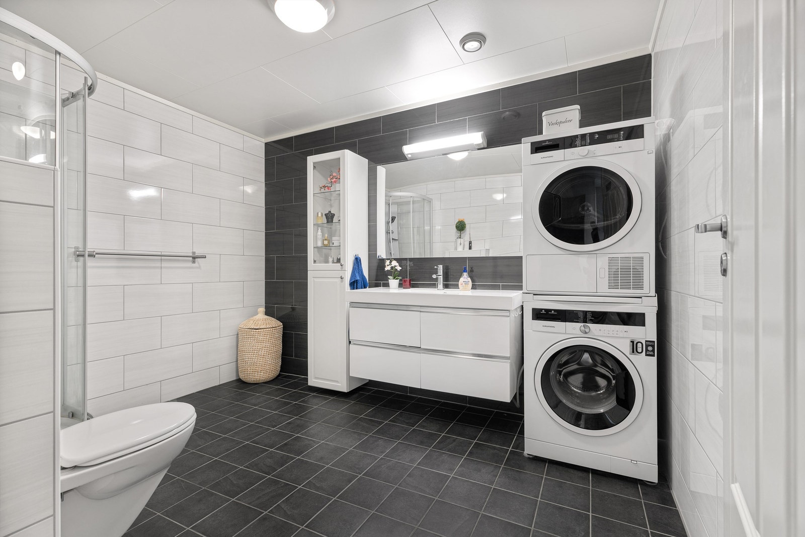 Romslig bad/wc/vaskerom med varmekabler som gir lun varme til nakne føtter. Moderne baderomsinnredning med stor servant. Vegghengt wc i hvit porselen. Opplegg for vaskemaskin og tørketrommel. Dusjkabinett montert i rommet. Galleribilde