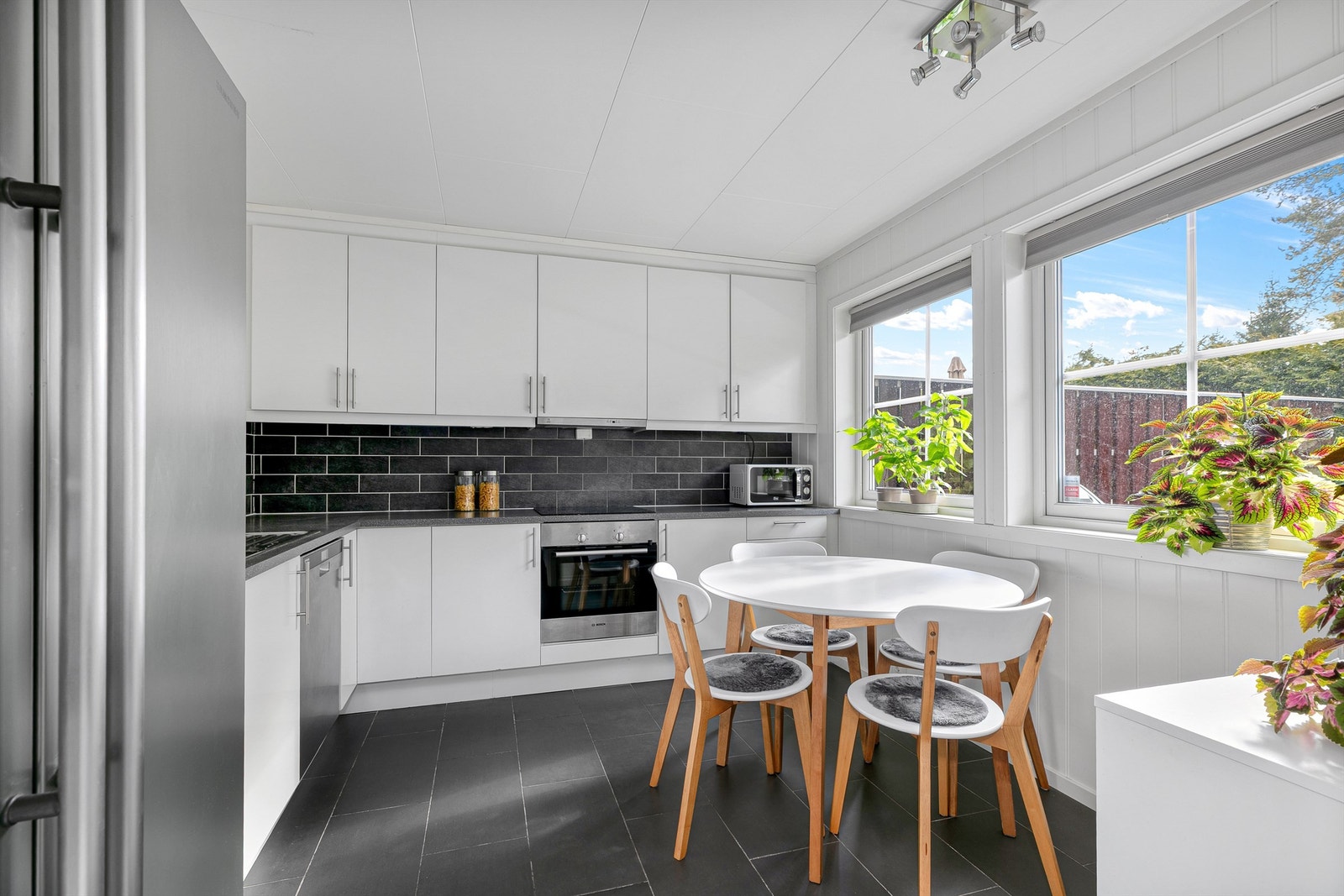 Moderne og stilrent kjøkken med flislagt vegg mellom sokkelskap og overskap som går til taket. Gode arbeidsflater på kjøkkenbenken som er lyssatt med lys under overskapene. Integrerte hvitevarer med komfyr, oppvaskmaskin. komfyrvakt montert. Galleribilde