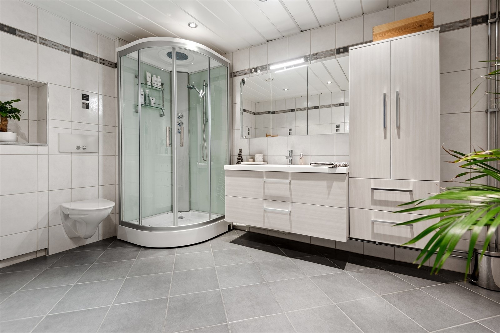 Badet er meget romslig og inneholder dusjkabinett, servant i baderomsmøblement med oppbevaring, overskap med speilfronter, og vegghengt toalett. Galleribilde