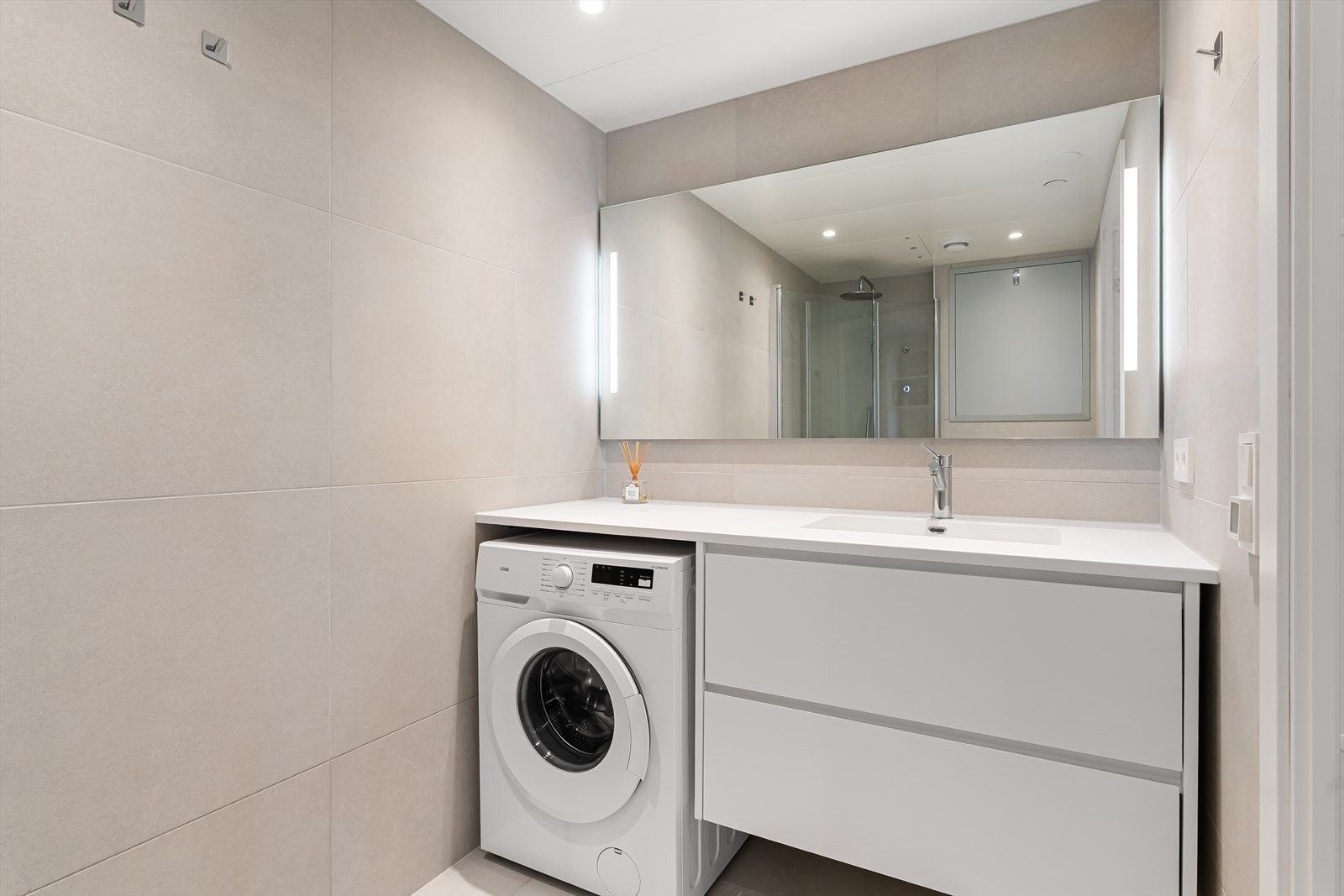 Badet har stort speil med innebygd lys, servant nedfeldt over skuffeløsning, vegghengt toalett, regnfallsdusj med innfellbare glassdører og opplegg for vaskemaskin Galleribilde