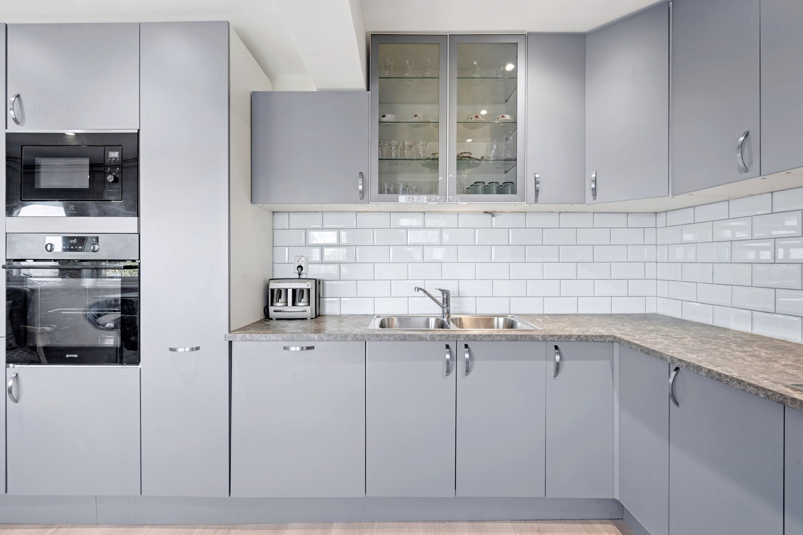 Kjøkkeninnredning med med grå matte fronter med takhøye skap, laminat benkeplater og fliser over benkeskap, rustfri oppvaskkum, ventilator med kullfilter og integrerte hvitevarer. Galleribilde