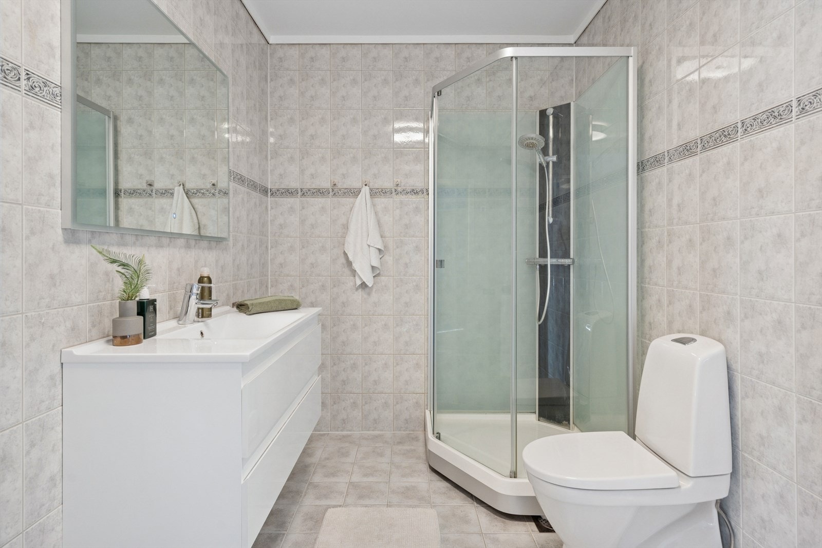 Innbydende helfliset bad/wc/vaskerom med varme i gulv. Rommet har toalett, innredning med servant, dusjkabinett, opplegg til vaskemaskin. Plastsluk. Ventilering via via balansert ventilasjon (avtrekksventil i tak samt luftespalte i dør). Galleribilde
