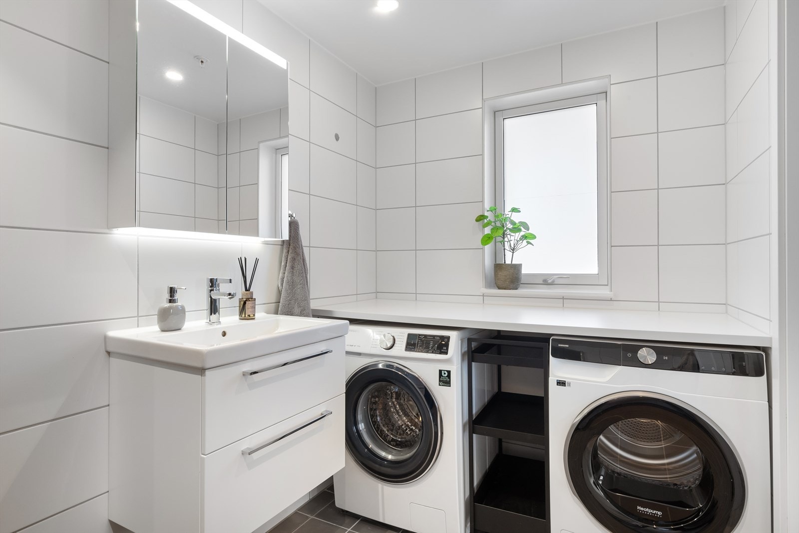 Badet er flislagt og gulvvarme. Badet er bygget opp med dusjhjørne med innfellbare glassdører, vegghengt servantinnredning, vegghengt toalett og speilskap, samt opplegg for vaskemaskin og tørketrommel under benk. Galleribilde