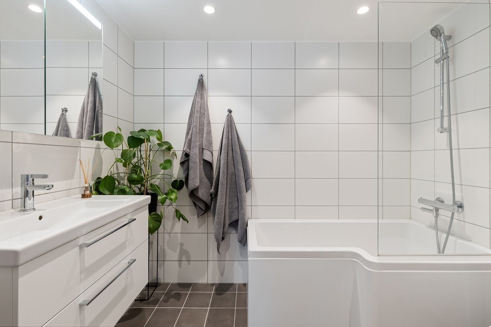 Lyst og delikat baderom med lyse fliser på veggene og mørke fliser på gulvet. På badet er det badekar med glassvegg, vegghengt servantinnredning og vegghengt toalett. Galleribilde