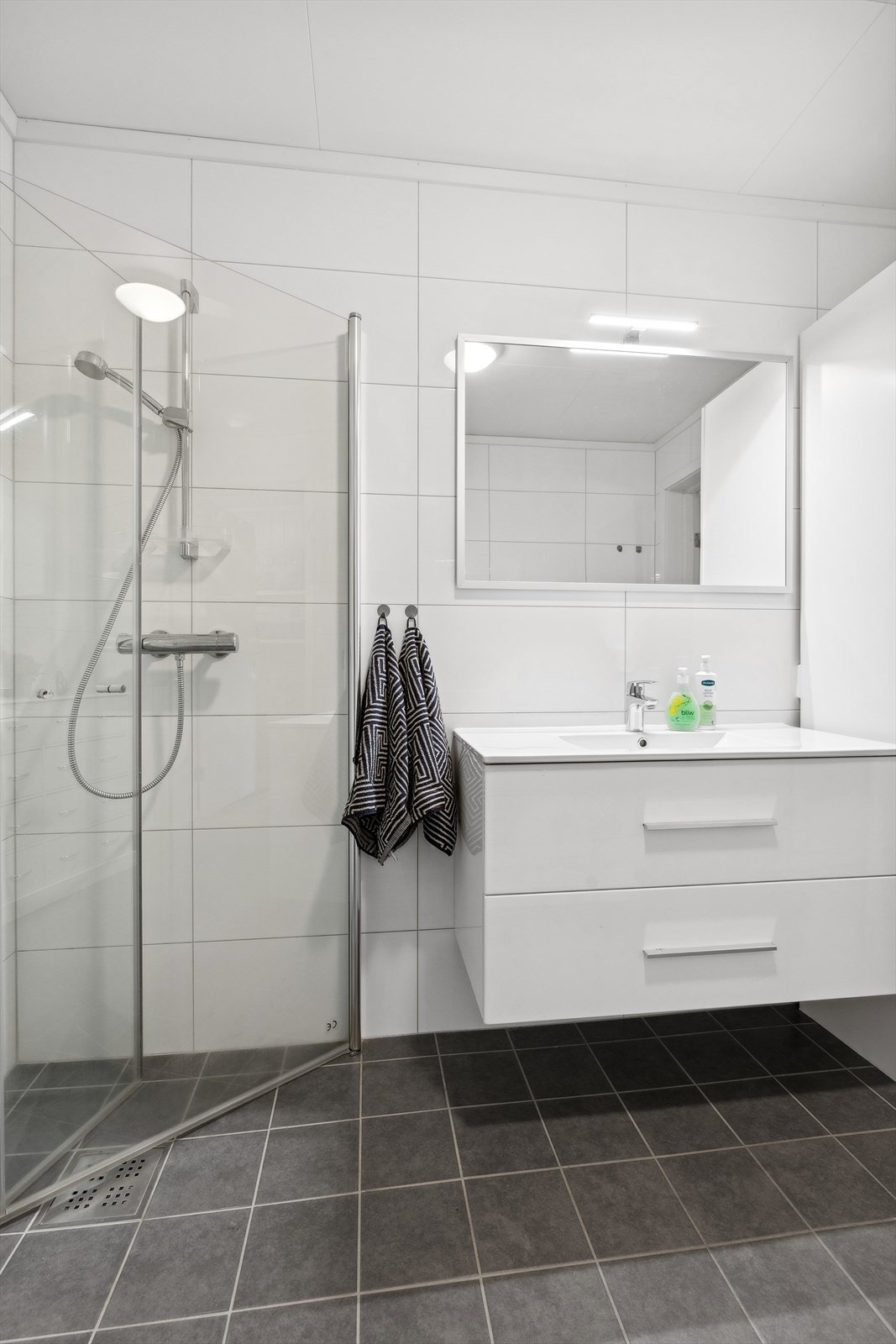 Badet er innredet med et veggmontert toalett, dusj med innfellbare dusjdører, baderomsinnredning med heldekkende servant og speil med overliggende belysning. Galleribilde