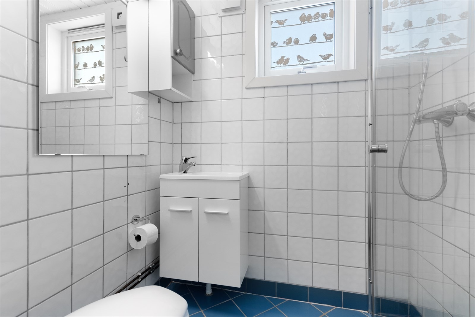 Badet har fliser på både vegger og gulv og er utstyrt med dusjhjørne, klosett og servant i innredning. Galleribilde