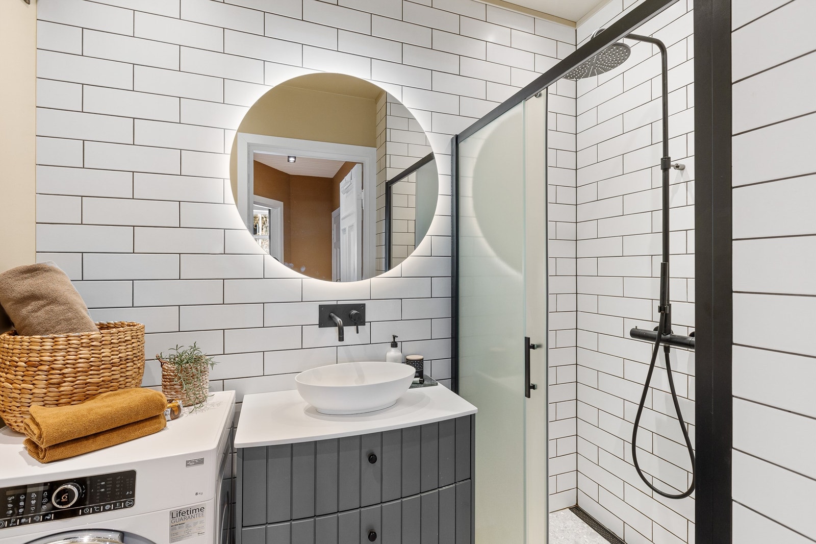 Badet har gulvstående servantinnredning med ovenpåliggende servant og speil med belysning, dusjnisje med glassdør og opplegg for vaskemaskin. Galleribilde