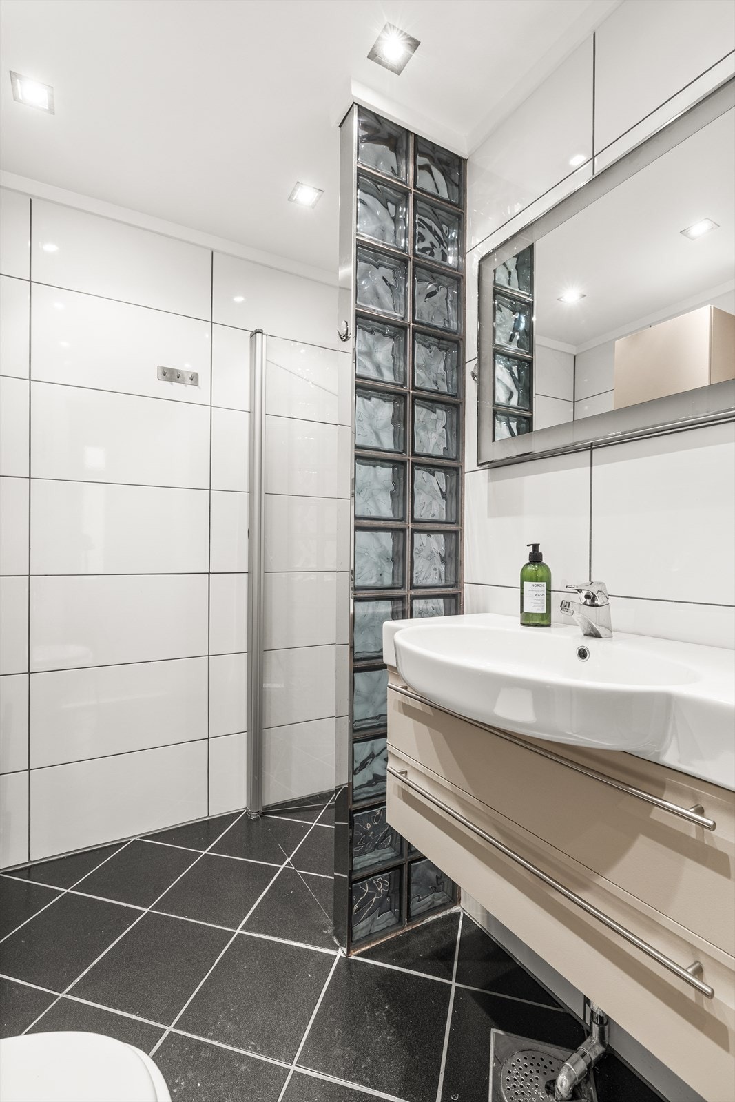 Delikat flislagt baderom innredet med dusjnisje med dusjvegg i glassbyggestein og dusjdør i glass, vegghengt toalett, servant med baderomsinnredning samt opplegg for vaskemaskin. Galleribilde