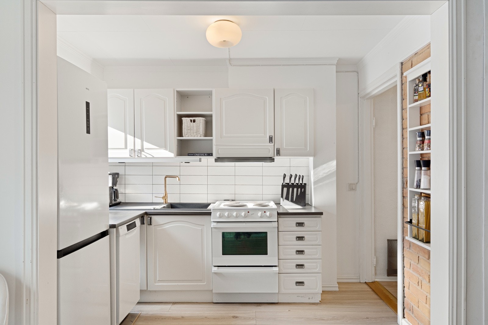 Kjøkkeninnredningen er montert i en funksjonell L-form, med hvite, profilerte trefiberfronter, gullfarget armatur og oppvaskkum nedfelt i en benkeplate av laminat. Galleribilde