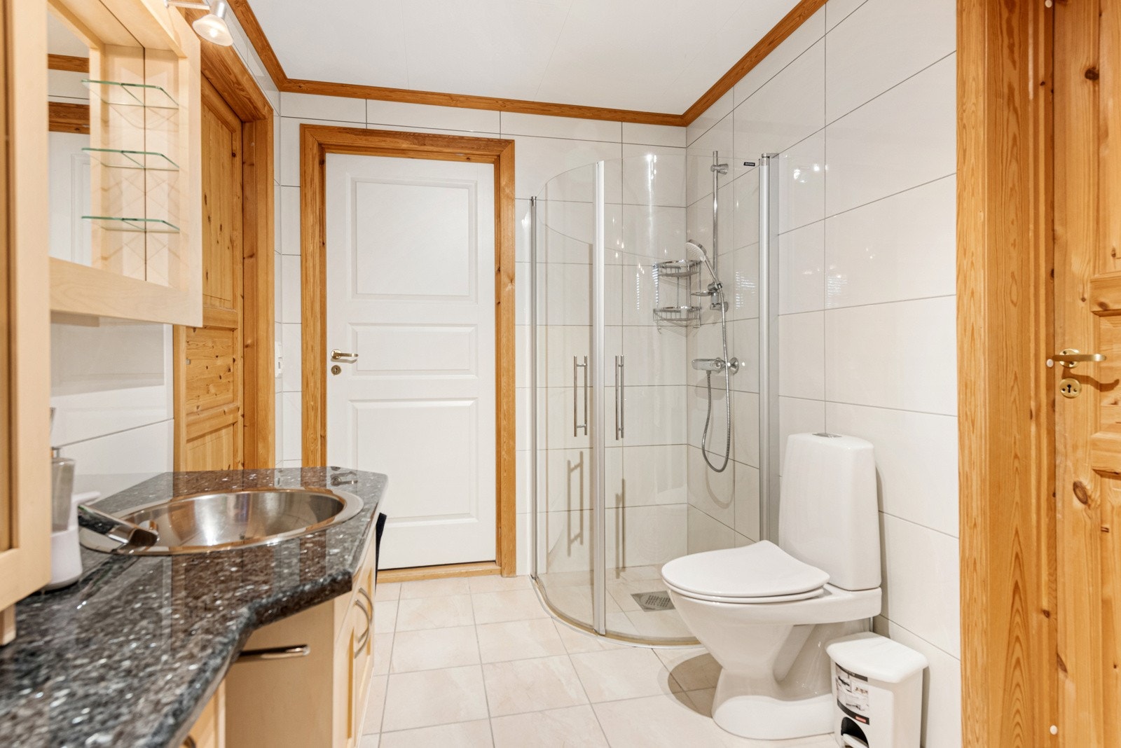 Baderomsinnredningen har profilerte fronter i en lys bjørkefarge og består av flere benkeskap, overskap med vitrine og to tilhørende speil med belysning. Det hele gjøres komplett med granittbenkeplate og to nedfelte servanter i stål. Galleribilde