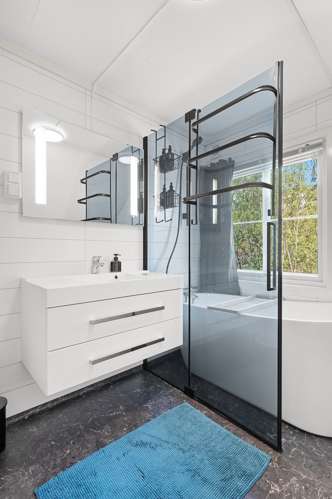 Badet inneholder enkel servant med underskaper, speil med automatisk lysfunksjon, moderne badekar med dusjvegg og et eldre toalett. Galleribilde