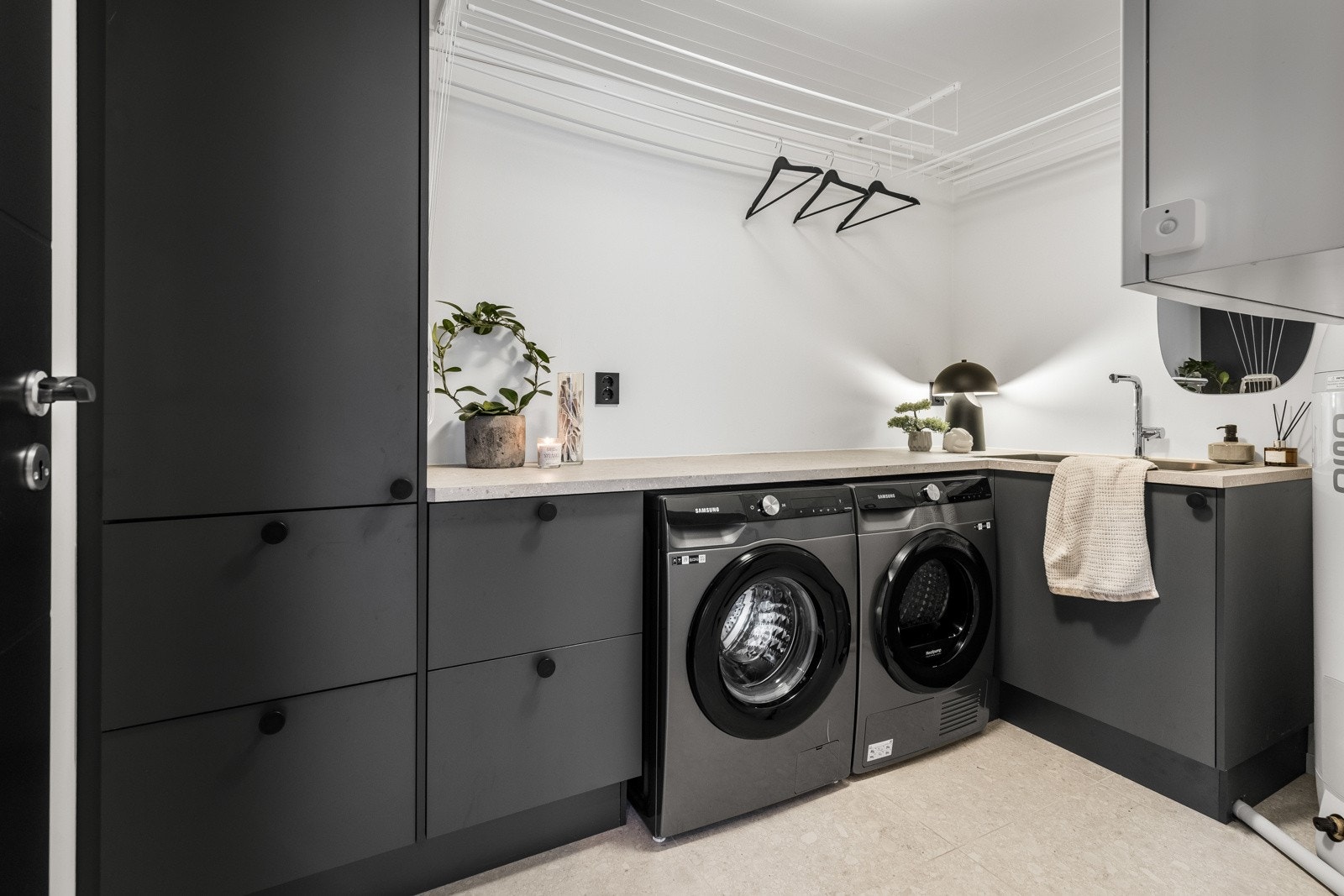 Vaskerommet har store, lysegrå gulvfliser med varme, og i en funksjonell L-form er en plassbygd innredning montert med smart oppbevaring. Skapseksjonen har sorte, glatte fronter og en stor arbeidsbenk med nedfelt utslagsvask. Galleribilde