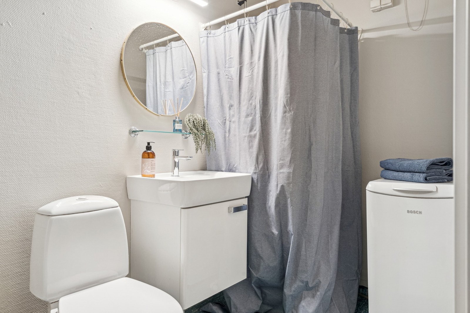 Badet er fra byggeår med belegg på gulv og tapet på vegger. Rommet er innredet med servant i møbel, dusjhjørnet, toalett og opplegg for vaskemaskin. Galleribilde