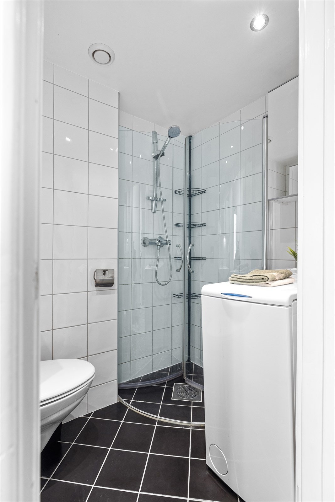 Badet inneholder dusjhjørne med innfellbare glassdører, vegghengt toalett og baderomsinnredning med servant, ett-greps blandebatteri, speil på vegg og skap. Galleribilde