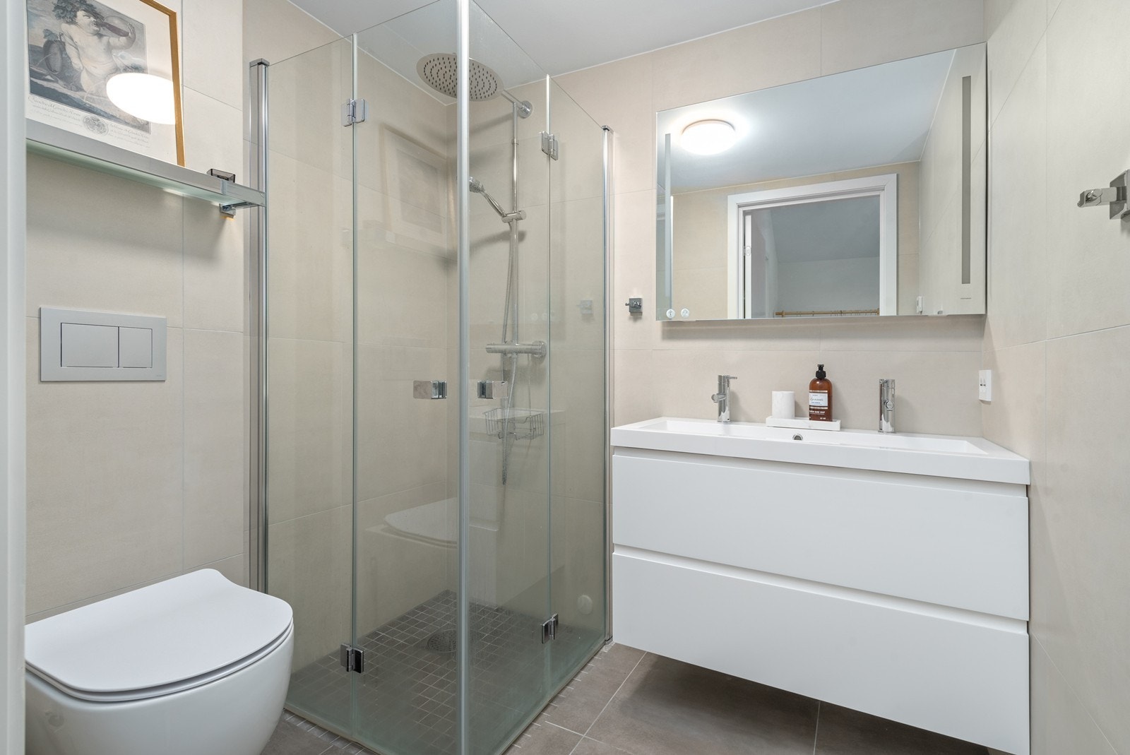 Flislagt bad med dusjhjørne, innredning med servant, skap og speil samt vegghengt toalett Galleribilde