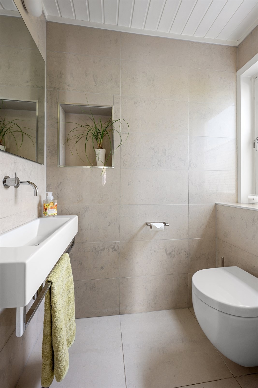 Gjestetoalett fra 2012 med flislagte overflater. Toalettet har et vegghengt wc og servant med ettgreps Vola blandebatteri. Over servanten er det speil med to veggmonterte lamper. Galleribilde