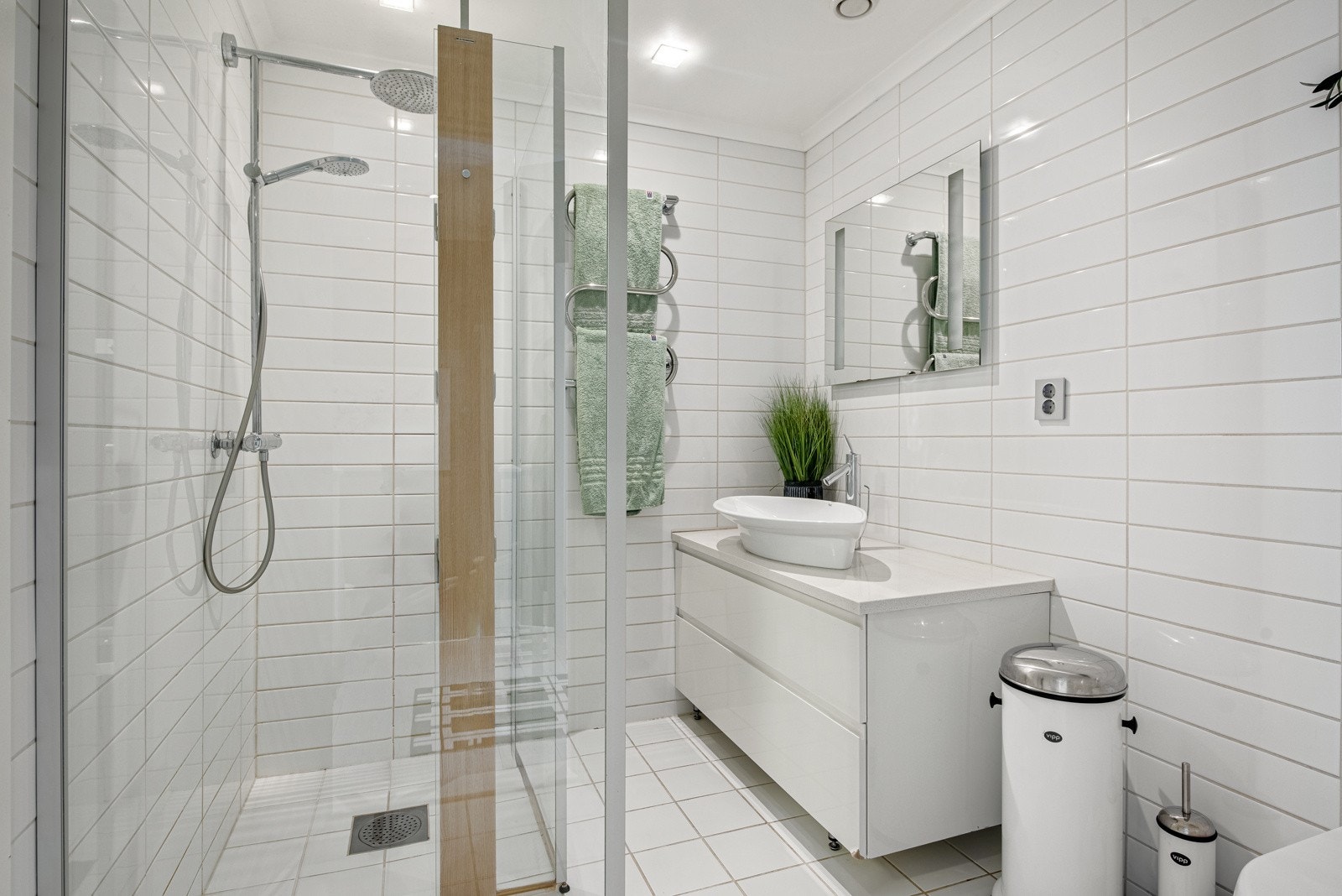 Baderomsinnredningen har skuffeseksjon med hvite, slette fronter, en stilig, toppmontert servant og speil med integrert belysning. I dusjhjørnet er det dusjvegger i glass og dusjgarnityr montert på vegg, med både hånddusj og regnfallsdusj. Galleribilde