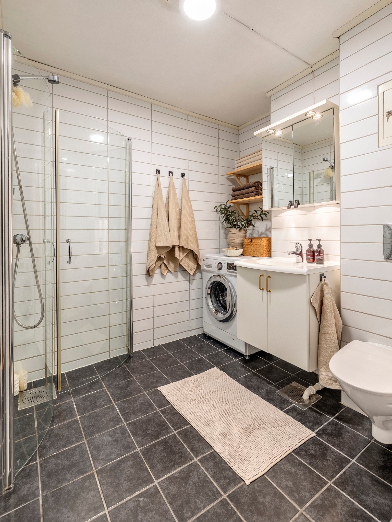 Badet er romslig med opplegg for vaskemaskin innerst i rommet, under trehvite vegghyller. Den øvrige innredningen består av en heldekkende servant på hvitt skap, speilskap med overlys, et veggmontert toalett og et dusjhjørne med innfellbare glassdører. Galleribilde