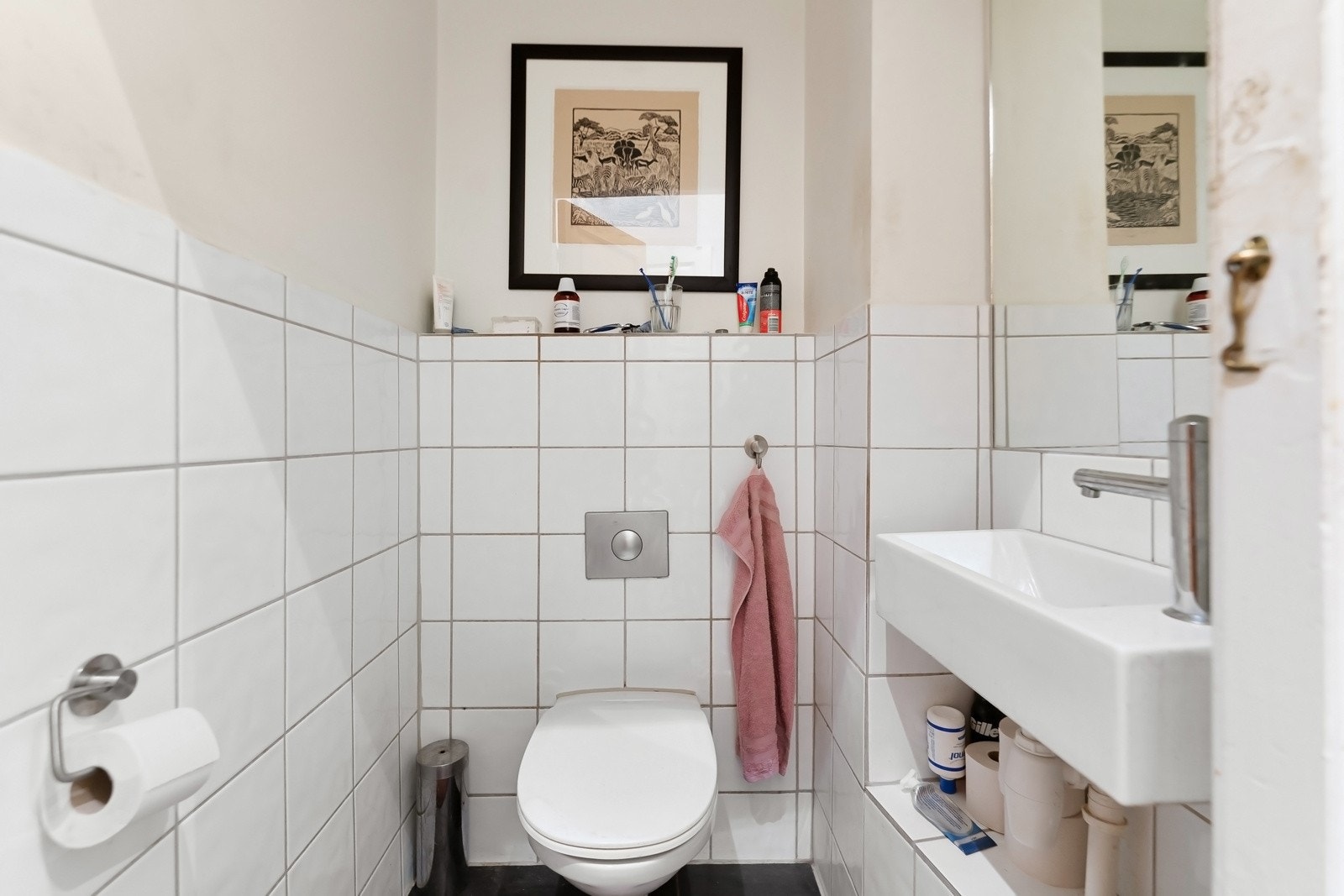 Toalettrommet er innredet med mørke fliser på gulvet, hvite fliser til brystningshøyde og malte vegger ellers, samt et veggmontert toalett, speil og servant. Galleribilde