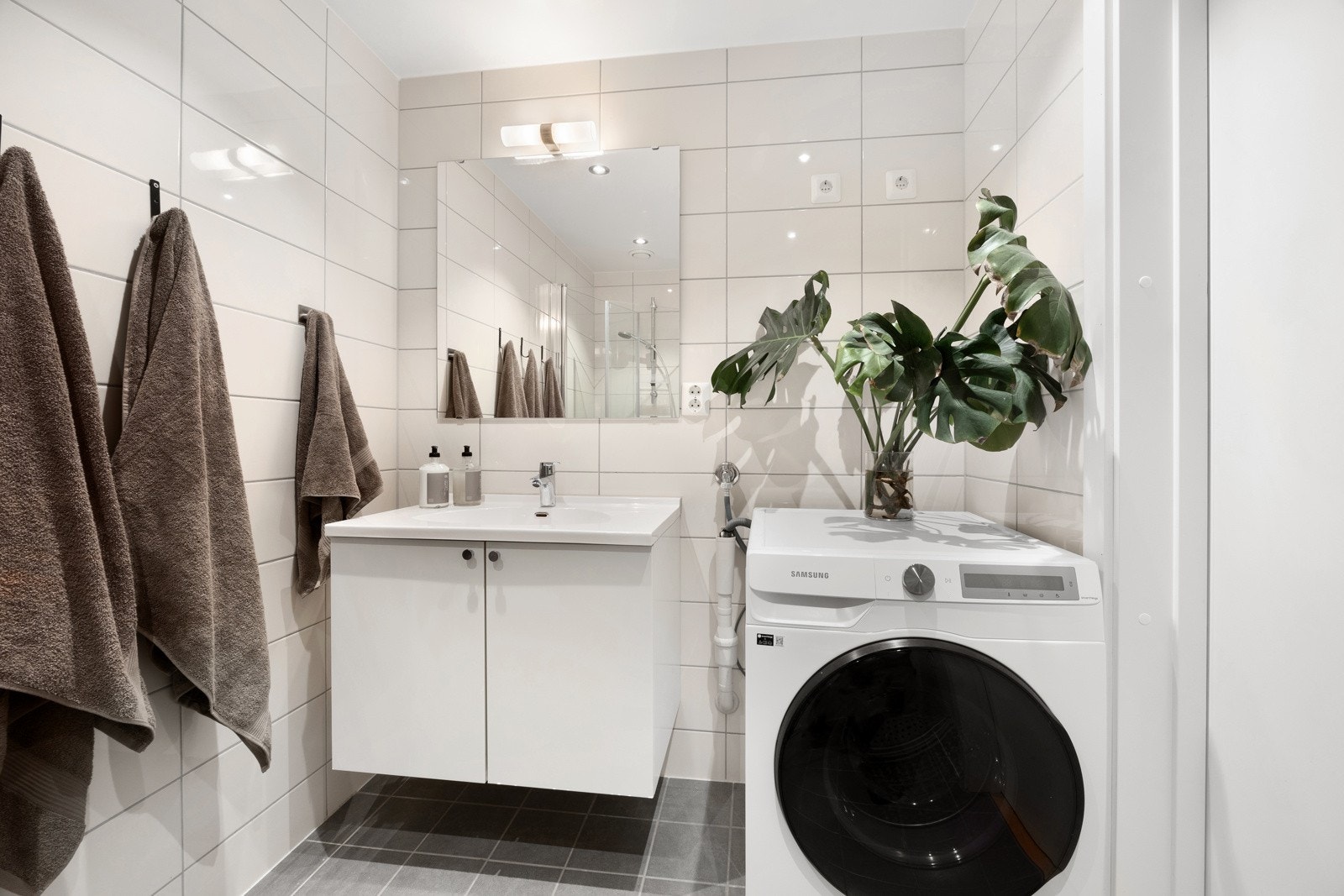 Baderommet inneholder vegghengt servantinnredning, ovenpåliggende servant, speil med belysning og stikkontakt over servant, og opplegg for vaskemaskin. Galleribilde