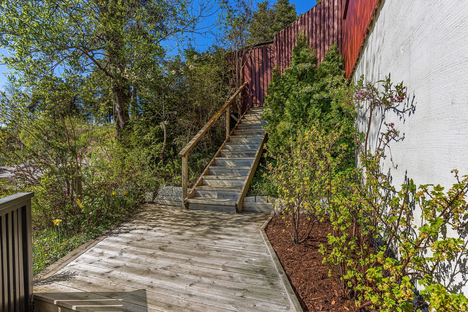 Terrassen på framsiden strekker seg rundt hushjørnet til gavlveggen der det er etablert en privat trapp opp til terrassen og hagen på baksiden. Området er beplantet med bl.a. bærbusker og morell- og ferskentre. Galleribilde