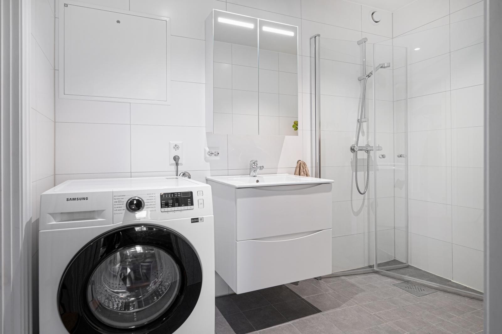 Badet har fliser på gulvet og baderomsplater på veggene. Her er dushjørne med innfellbare glassdører og opplegg for vaskemaskin. Galleribilde