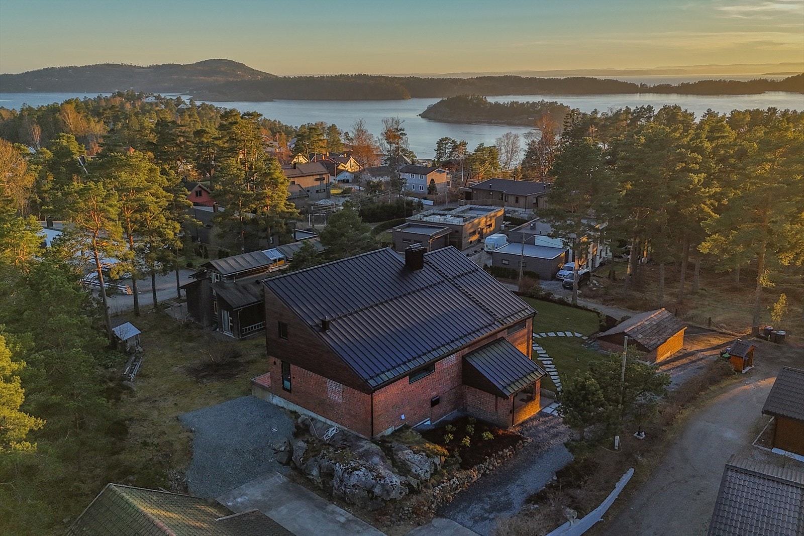 Tømmeråsen er et moderne hus med en distinkt arkitektonisk stil. Huset har to synlige fasader, én med mørk brun trepanel og den andre med rød murstein. Galleribilde