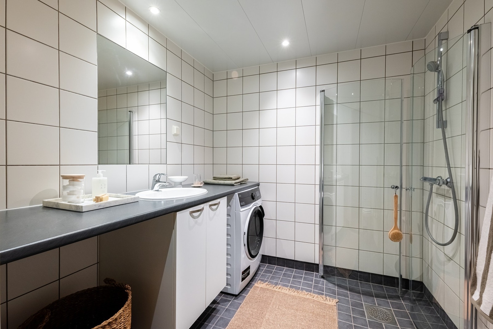 Lyst, flislagt bad med varme i gulv og downlights i himling. Praktisk nisje for plassering av vaskemaskin og tørketrommel under benk. Galleribilde