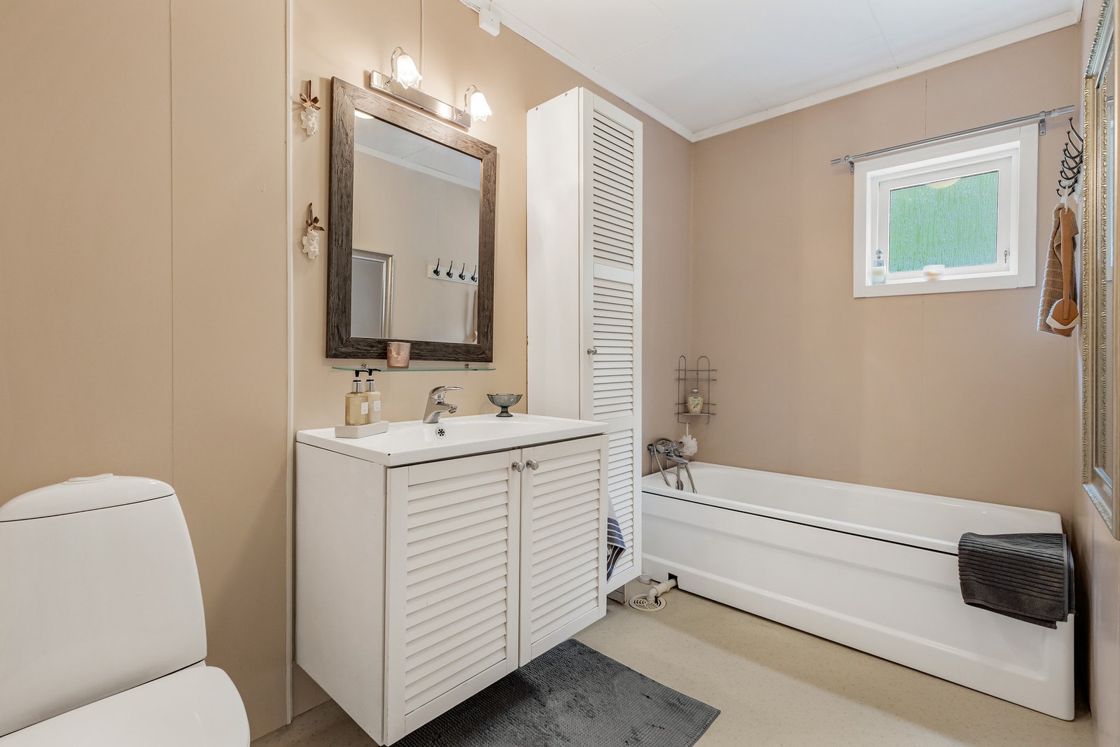 Badet er utstyrt med et gulvstående toalett og vegghengt servantskap med heldekkende servant samt badekar med togreps blandebatteri Galleribilde