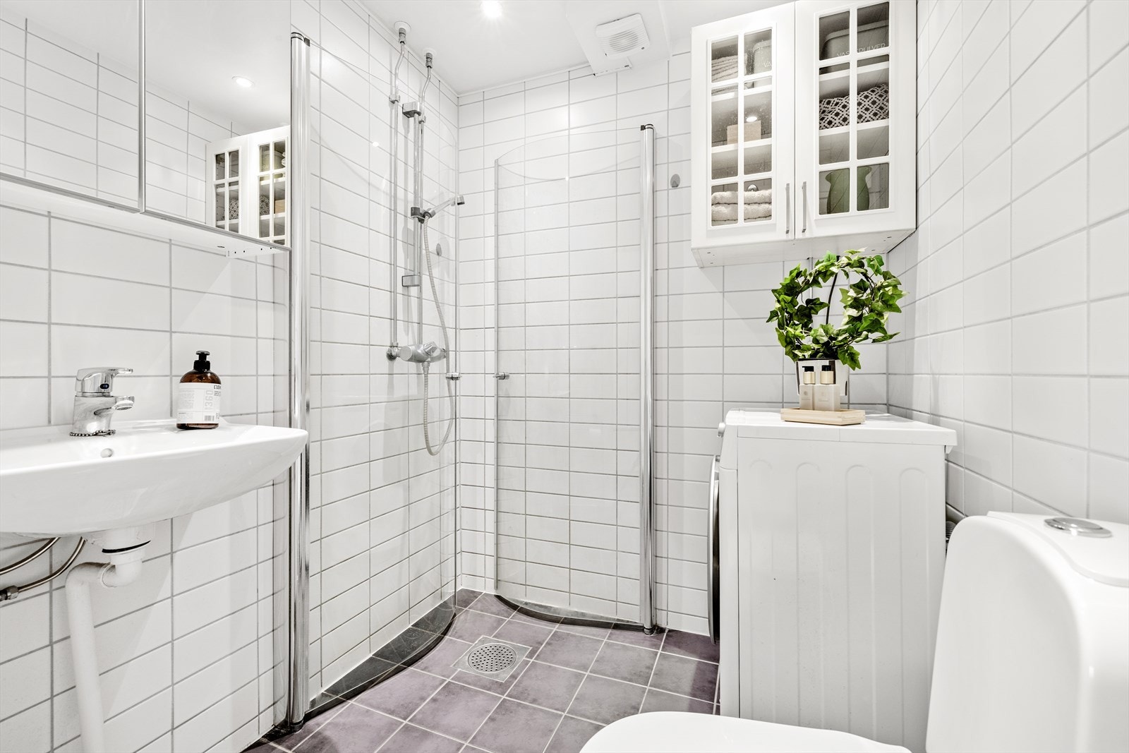 Baderom med fliser på gulv og vegg. Rommet er innredet med gulvmontert klosett, nedsenket vask med innredning og speil, dusjnisje med glassdører og opplegg til vaskemaskin og tørketrommel. Badet ble renovert i regi av borettslaget i 2007. Galleribilde