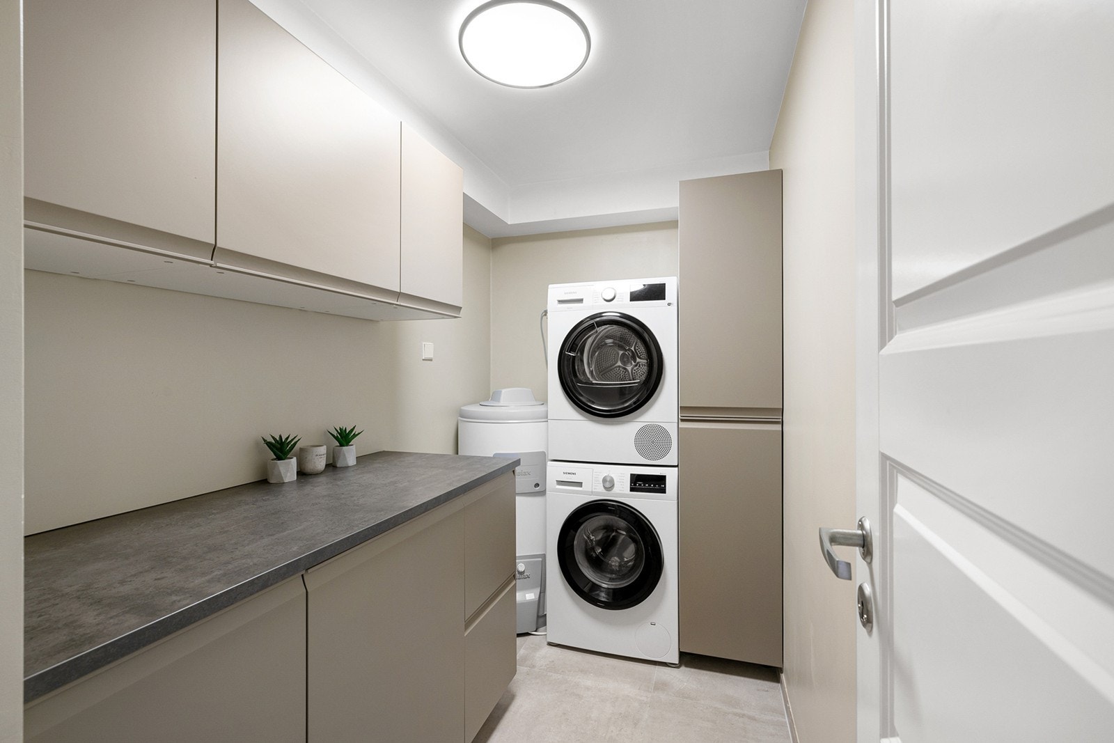 Et nytt og praktisk vaskerom med fliser på gulv med gulvvarme og malte veggflater. Innredning fra 2023. Det er opplegg for vaskemaskin og tørketrommel. Galleribilde