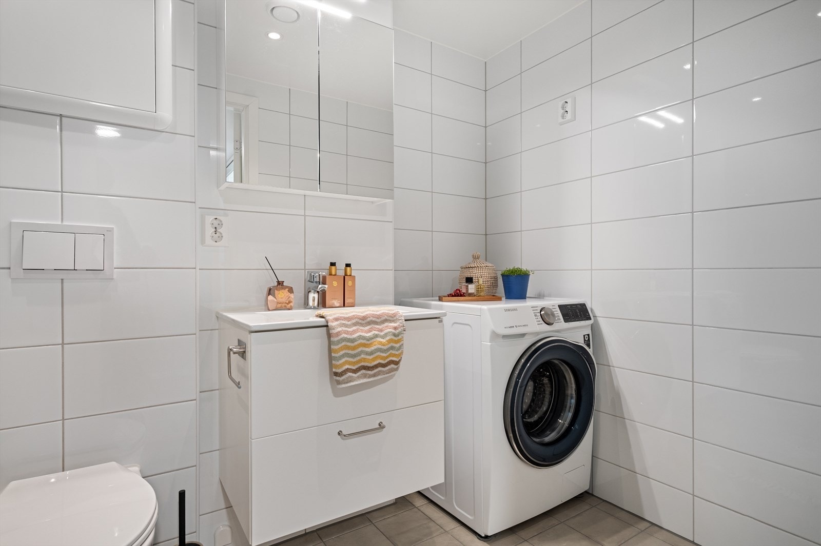 Badet er innredet med servantmbøler, overskap, vegghengt toalett, og dusjhjørne. Det er for øvrig varmekabler i gulv og opplegg for vaskemaskin og tørketrommel. Galleribilde